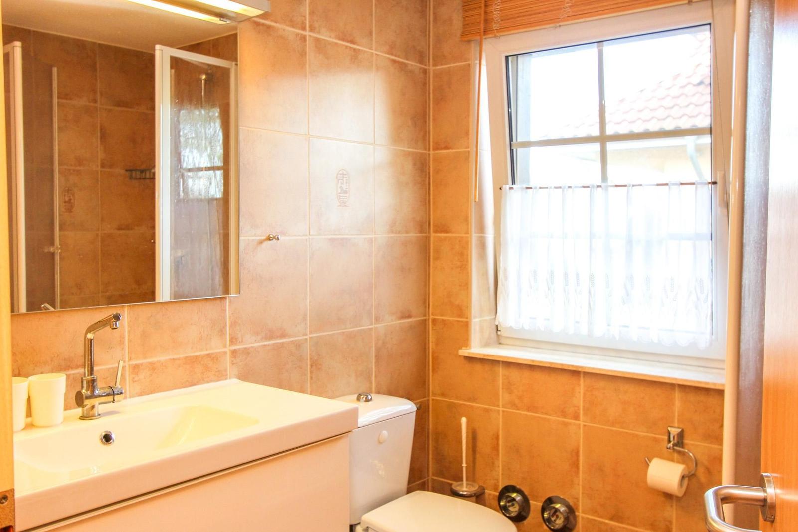 Badezimmer mit Waschbecken, Toilette und Dusche. Fenster mit weißen Vorhängen.