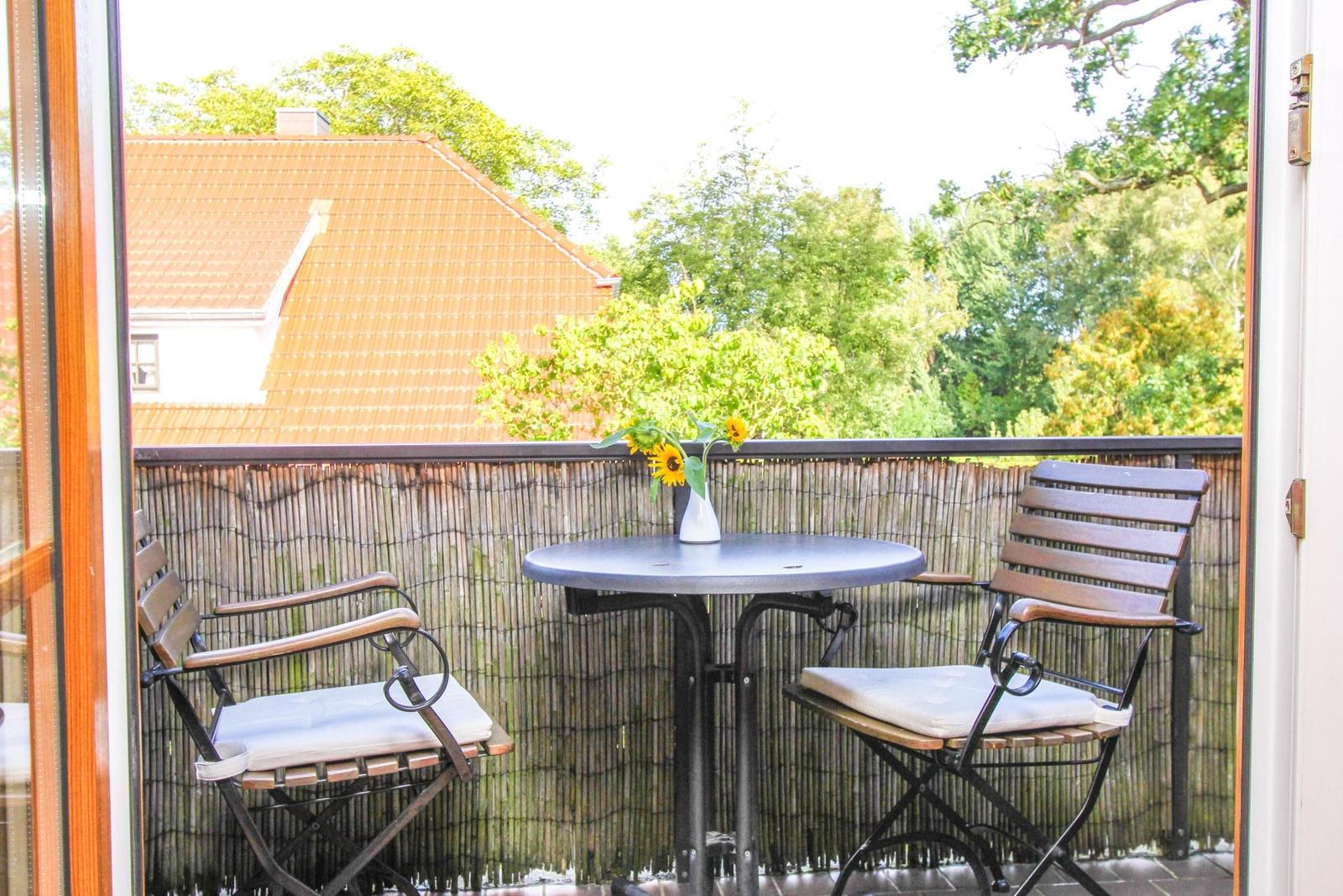 Balkon mit Tisch und zwei Stühlen, Sonnenblumen im Vasen, Blick auf Dach und Bäume.