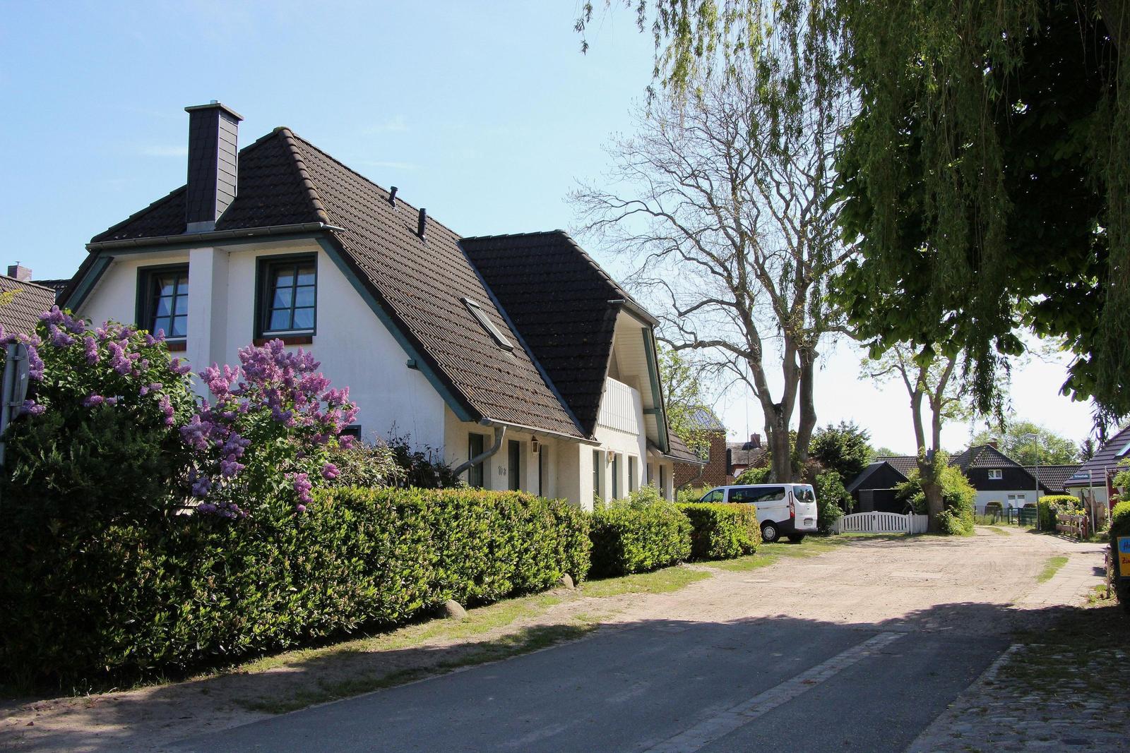 Wohnhaus mit dunklem Dach und grüner Hecke, umgeben von Bäumen und blühenden Sträuchern.