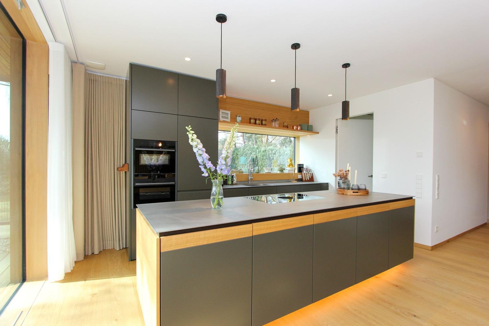 Moderne Küche mit Insel, Holzakzenten und integriertem Backofen. Große Fensterfront mit Blick nach draußen.