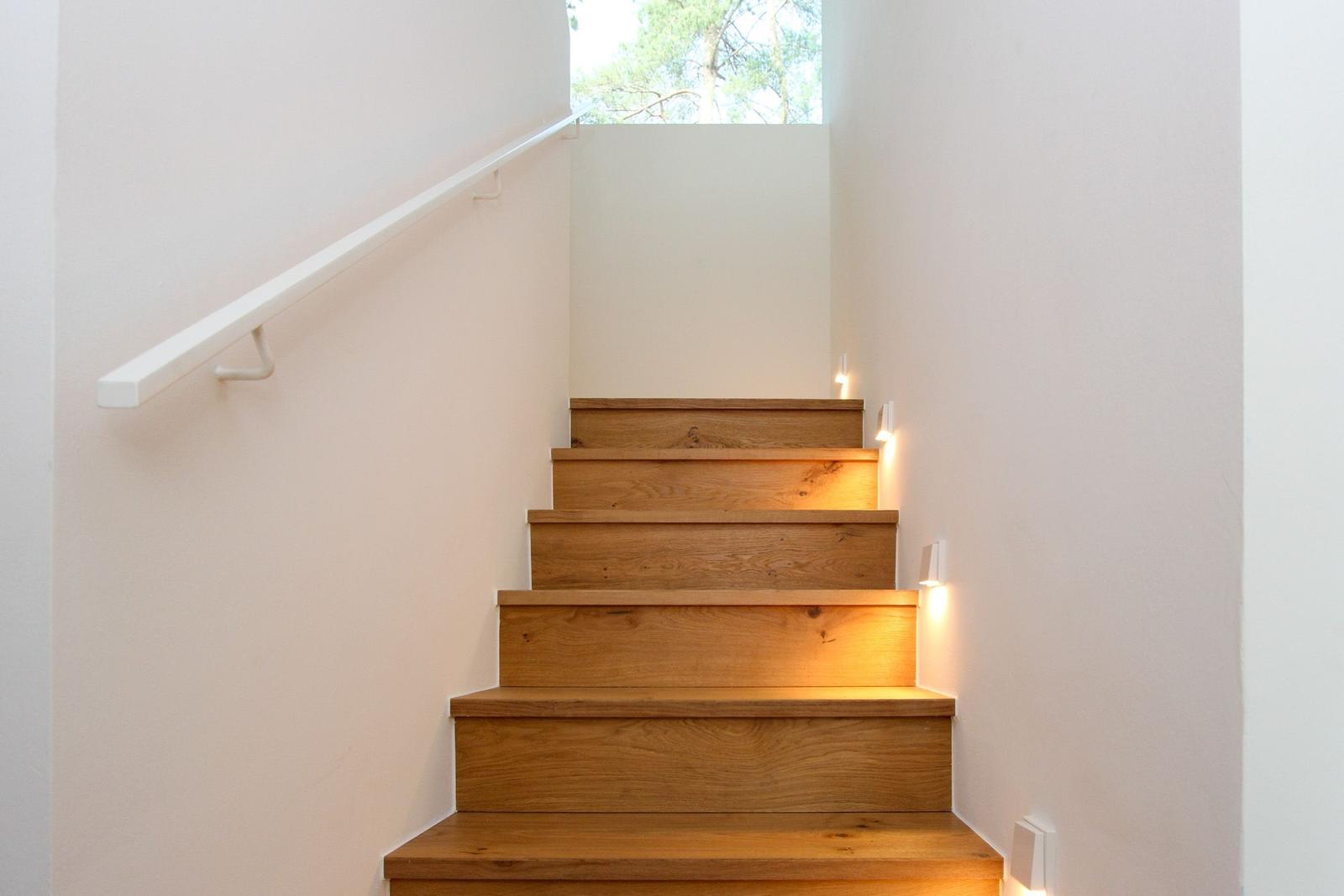 Holzgetreppe mit weißer Handlauf und Wandbeleuchtung, Blick auf Baum durch Öffnung.