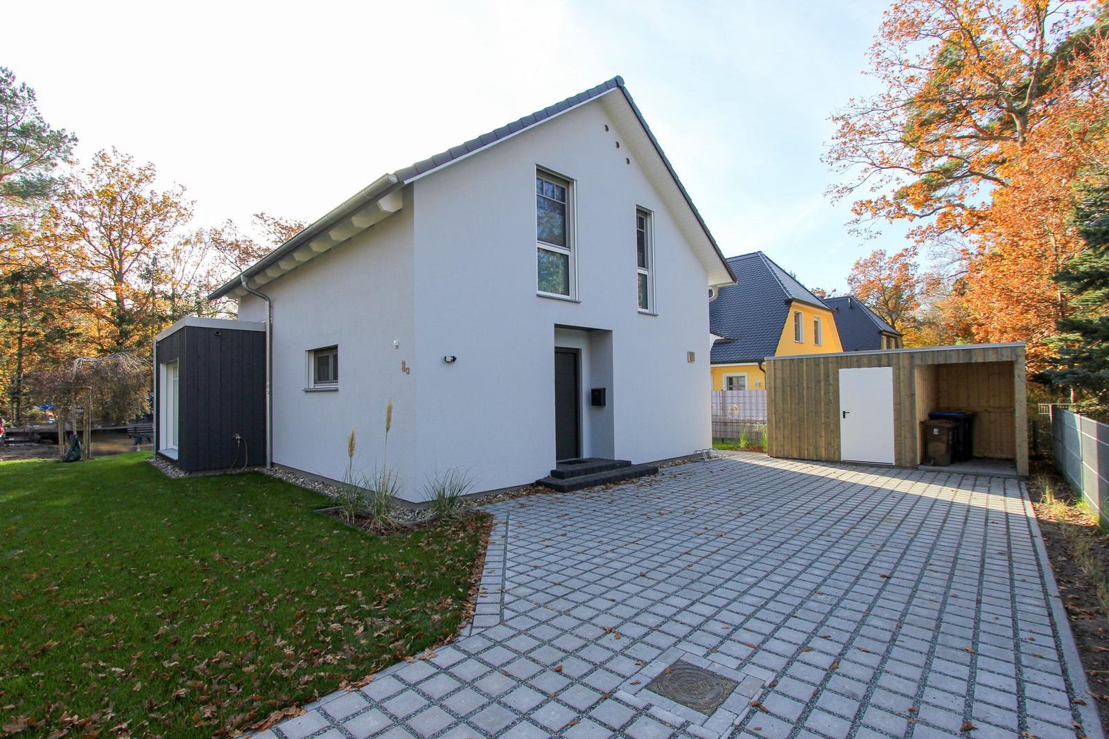 Moderne weiße Villa mit gepflastertem Hof und grünem Garten. Umgeben von Bäumen mit Herbstlaub.