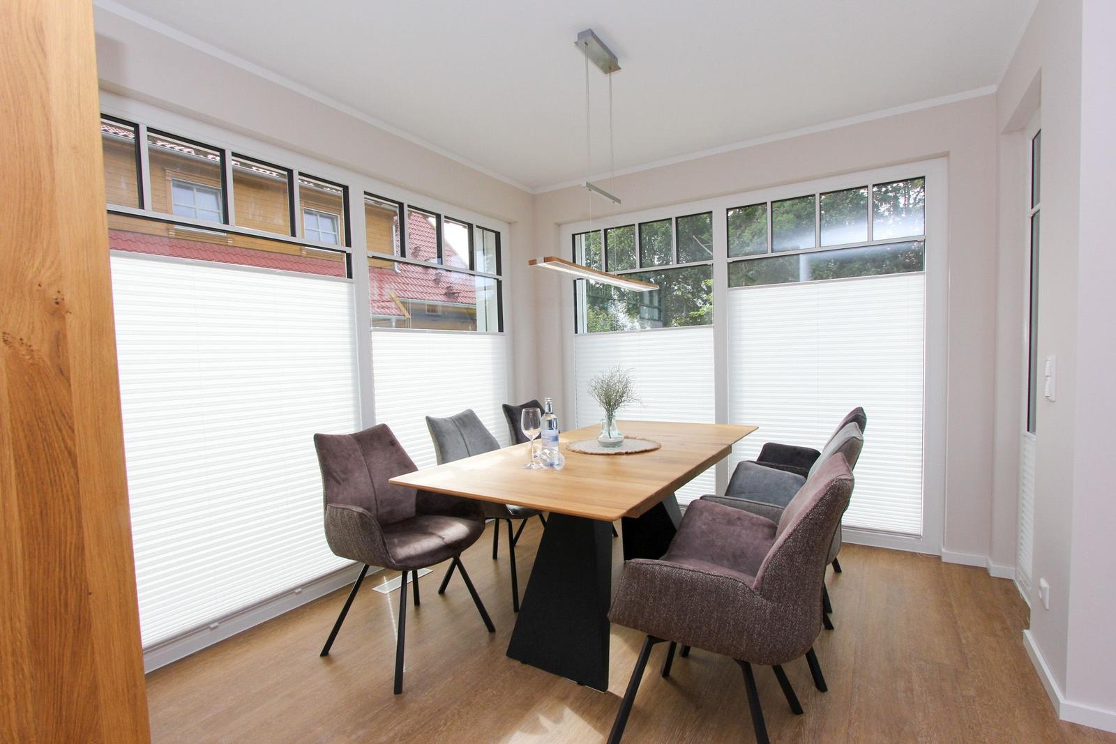 Esszimmer mit Holztisch, Stühlen und großen Fenstern mit Jalousien.
