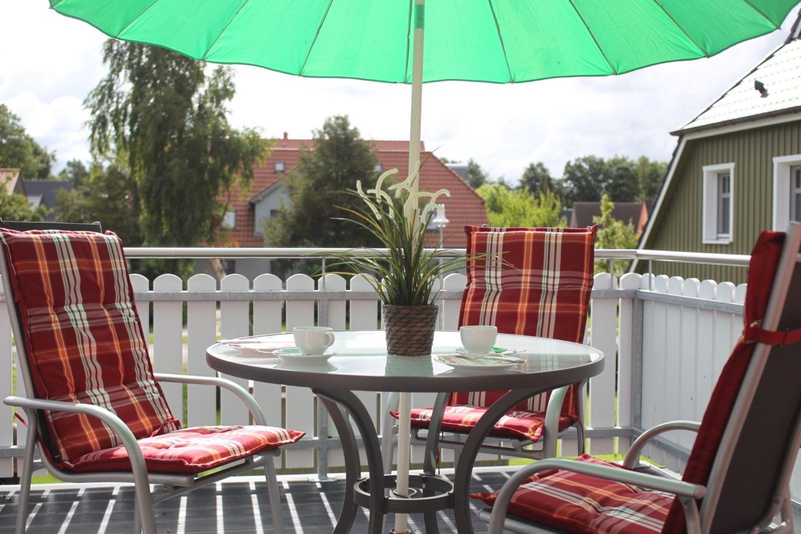 Terrasse mit Tisch, drei Stühlen und grünem Sonnenschirm. Hintergrund: weiße Zaun und Häuser.