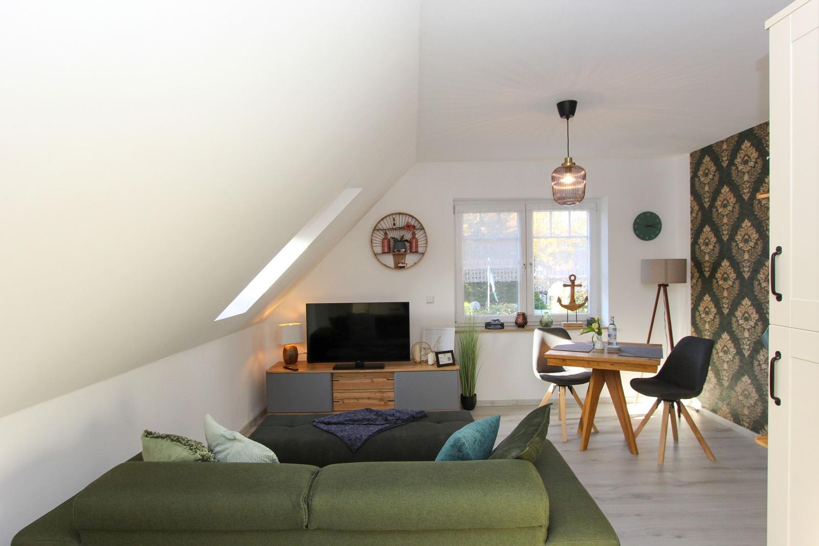 Wohn- und Essbereich mit Sofa, TV und Tisch. Dachschräge, Fenster und Wand mit Muster.