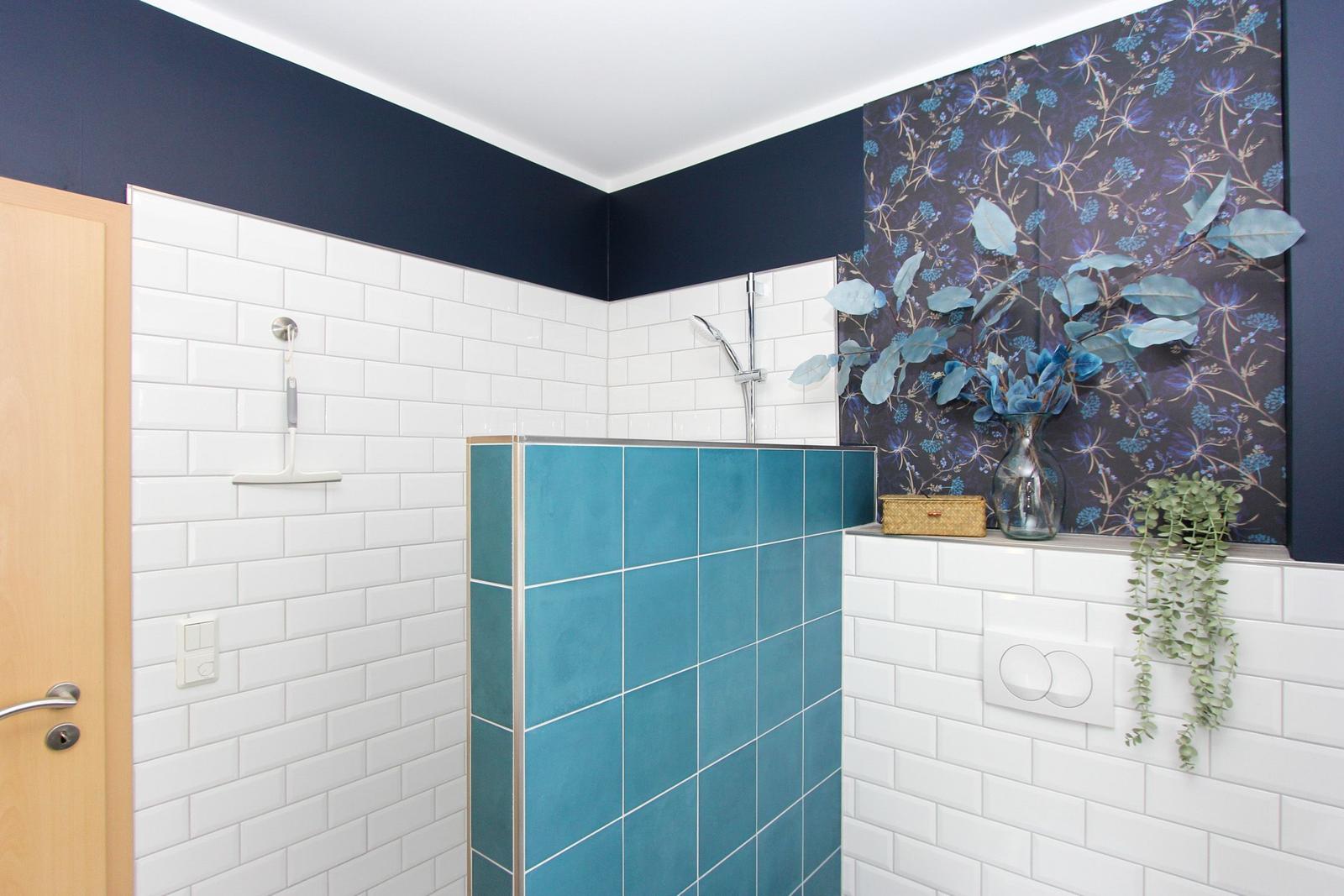 Bad mit Dusche, blauen Fliesen und floraler Tapete. Holztür und Pflanzen sind sichtbar.