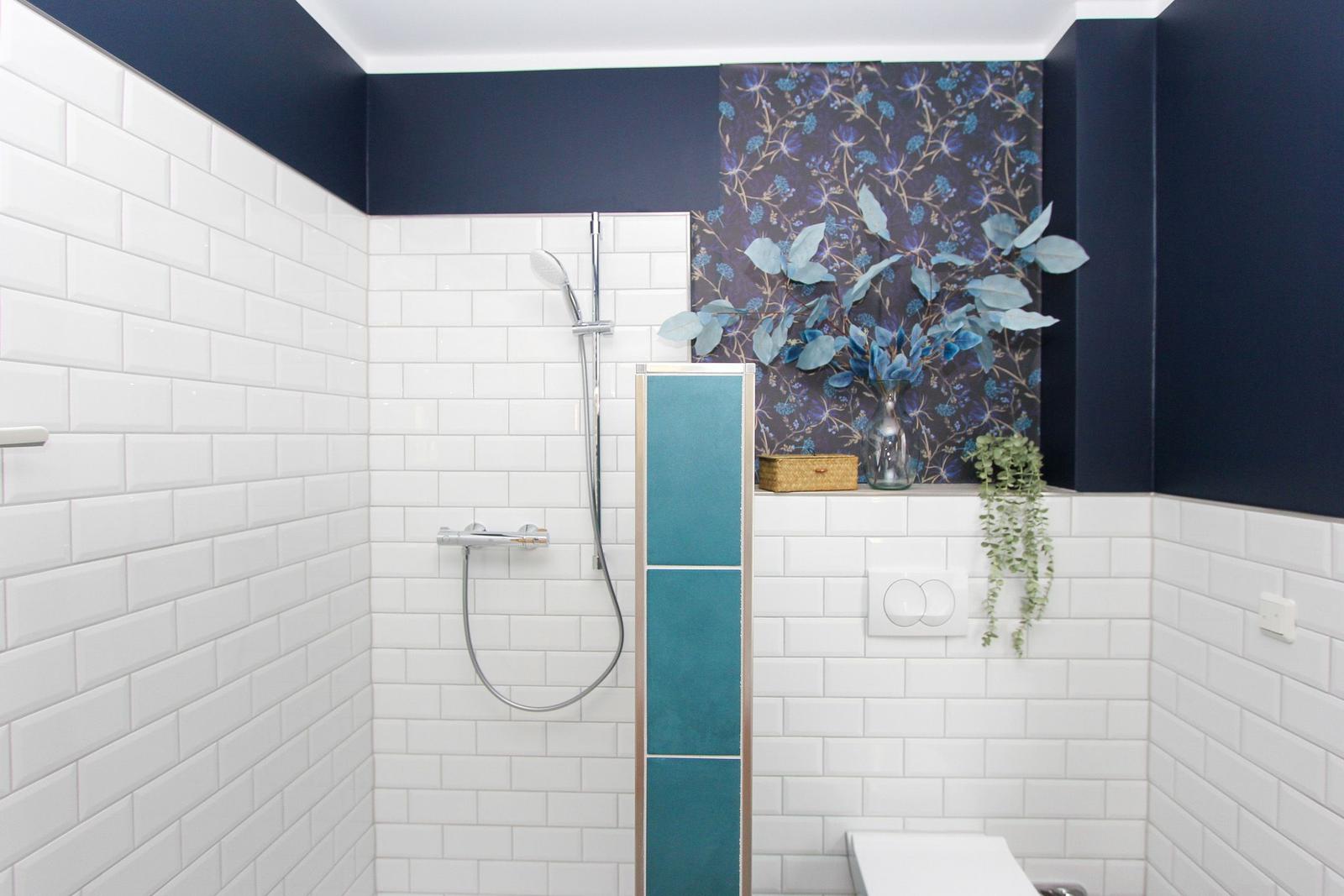 Duschbereich mit weißen Fliesen, blauem Wandpaneel und floraler Tapete. Dusche mit Handbrause und Toilette.