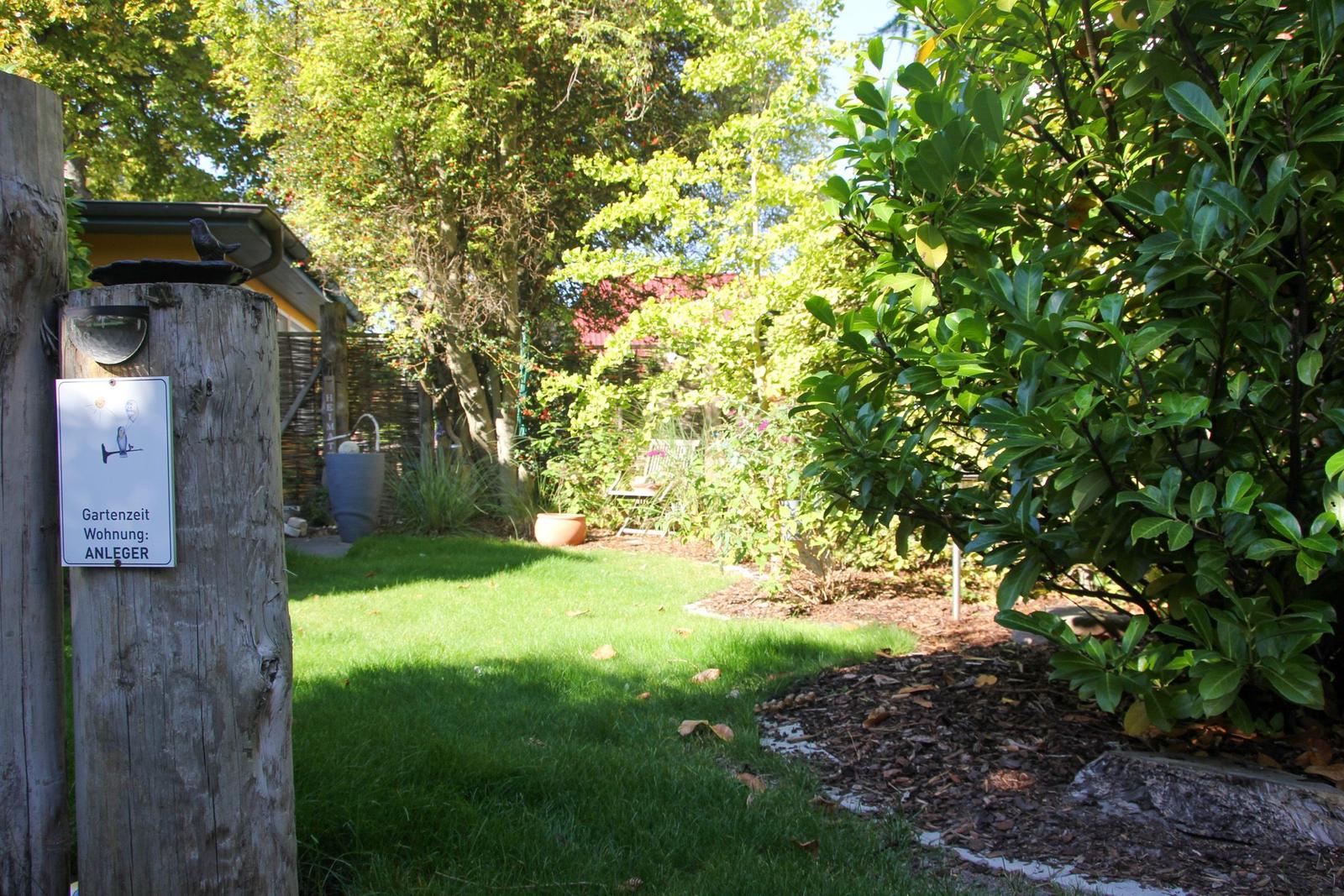 Garten mit Rasen, Sträuchern und Schild. Im Hintergrund ist ein Gartenhaus sichtbar.
