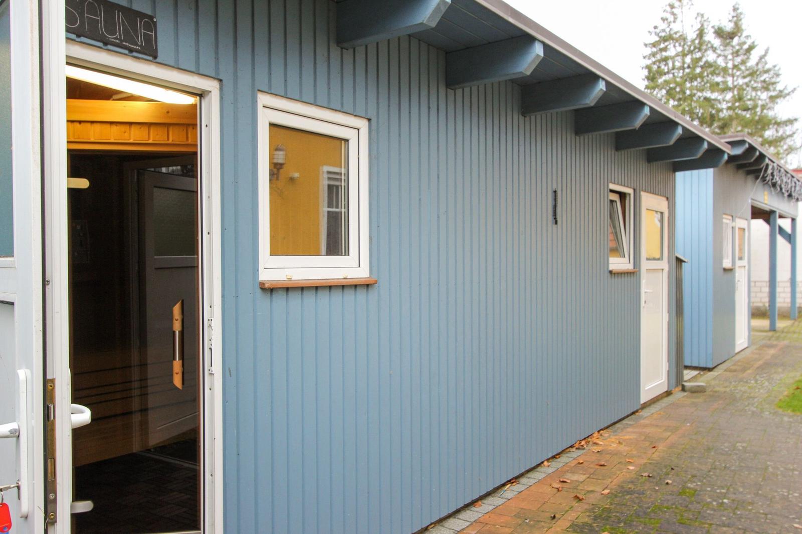 Sauna-Eingang mit offener Tür und Holzwand im Inneren. Außenwand aus blauen Wellblechplatten.