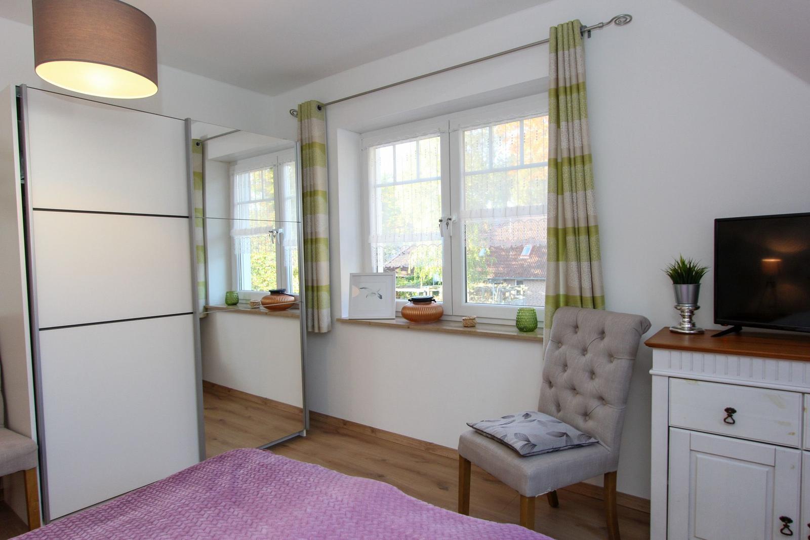 Schlafzimmer mit Bett, Kleiderschrank, Sessel und TV. Fenster mit Vorhängen.