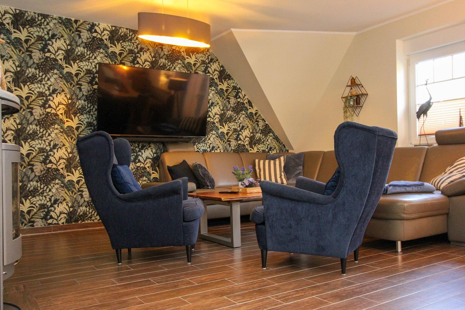 Wohnzimmer mit Sofa, zwei blauen Sesseln und Fernseher an floraler Wand.