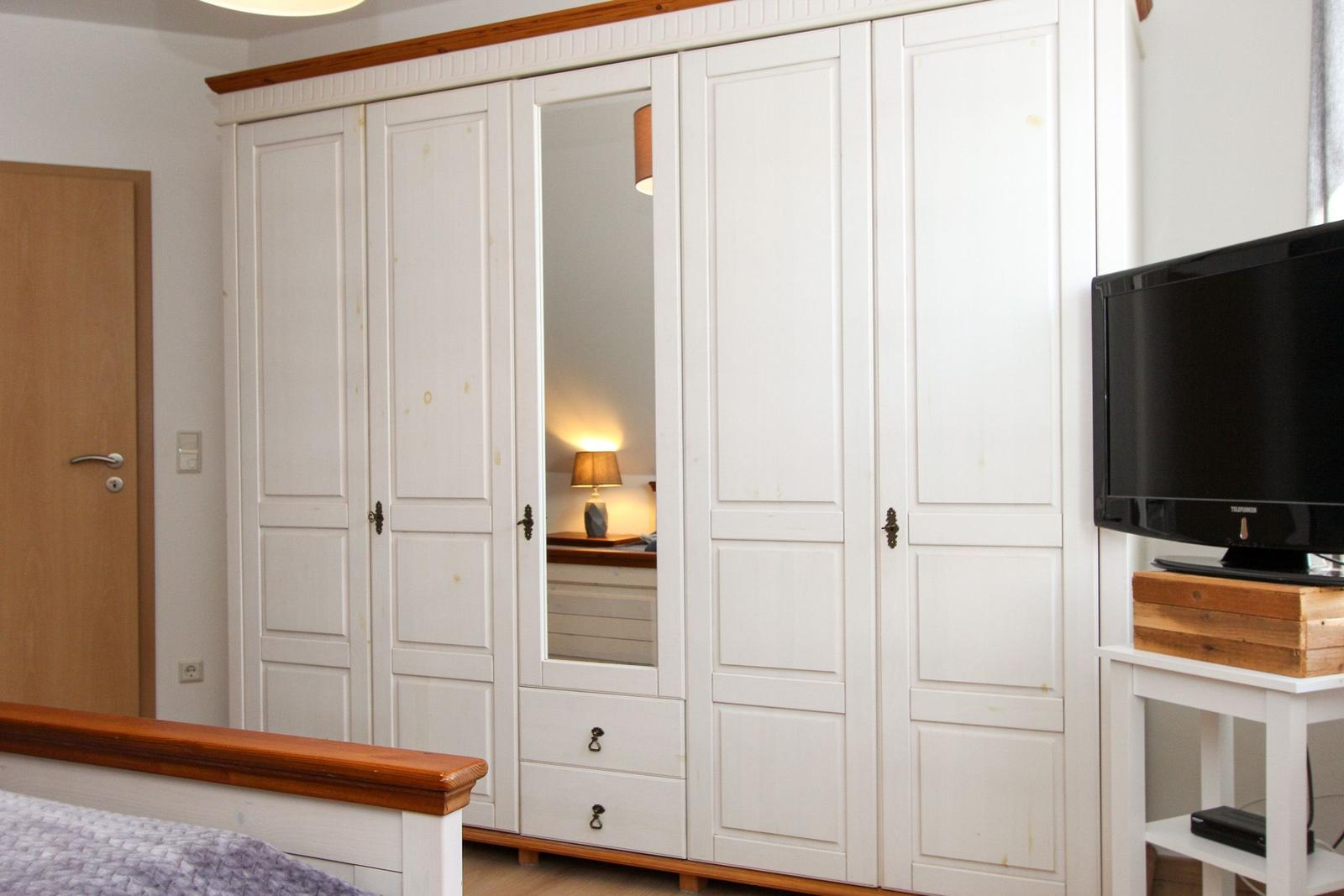 Schlafzimmer mit weißem Schrank, Spiegeltür und Fernseher auf Holzregal.