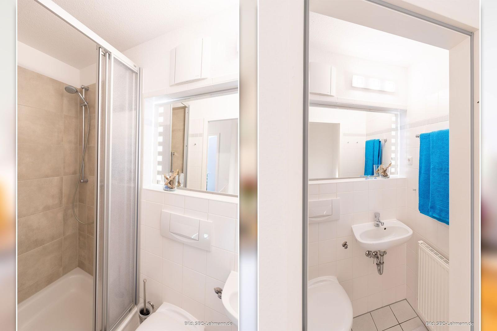 Badezimmer mit Dusche, Waschbecken und Toilette. Spiegel und blaue Handtücher sichtbar.