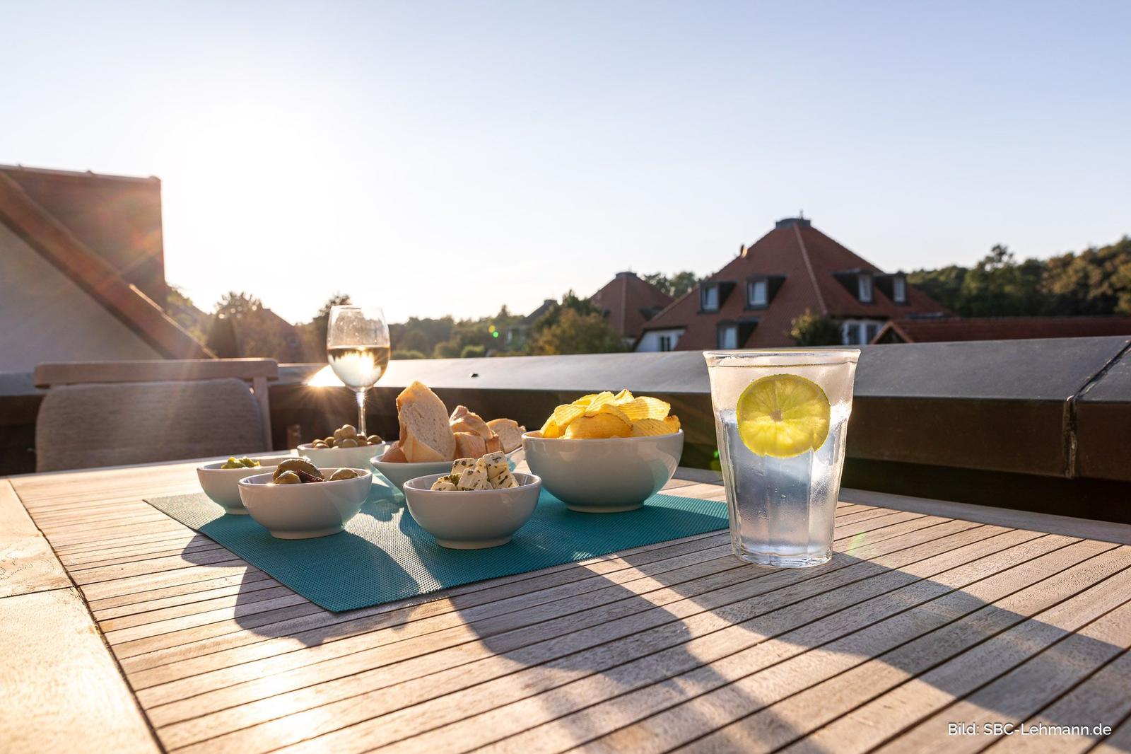 Auf der Terrasse steht ein Tisch mit Speisen und Getränken. Im Hintergrund sind Häuser und Bäume zu sehen.