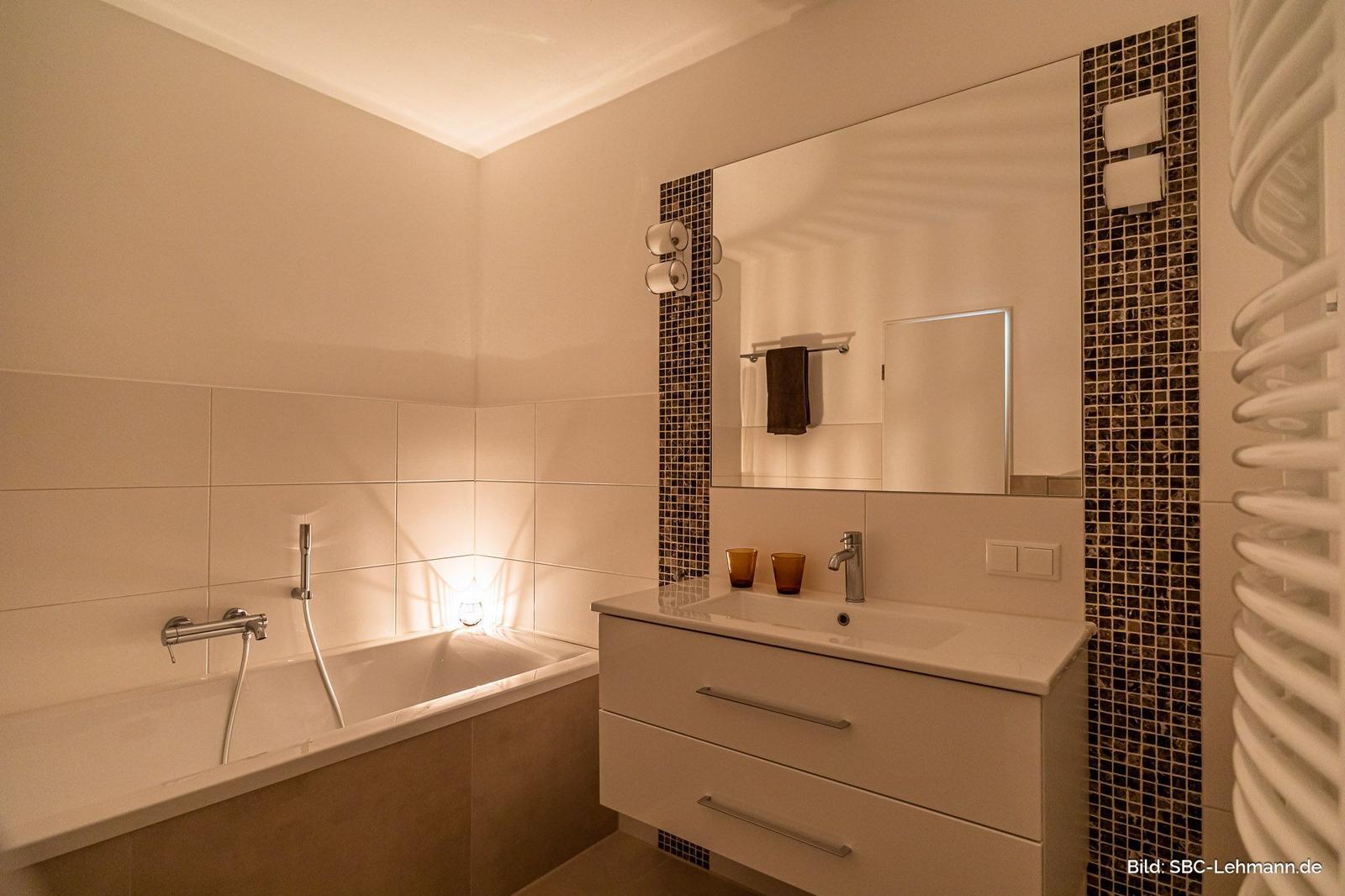 Badezimmer mit Badewanne, Waschtisch und Spiegel mit Mosaikverkleidung.