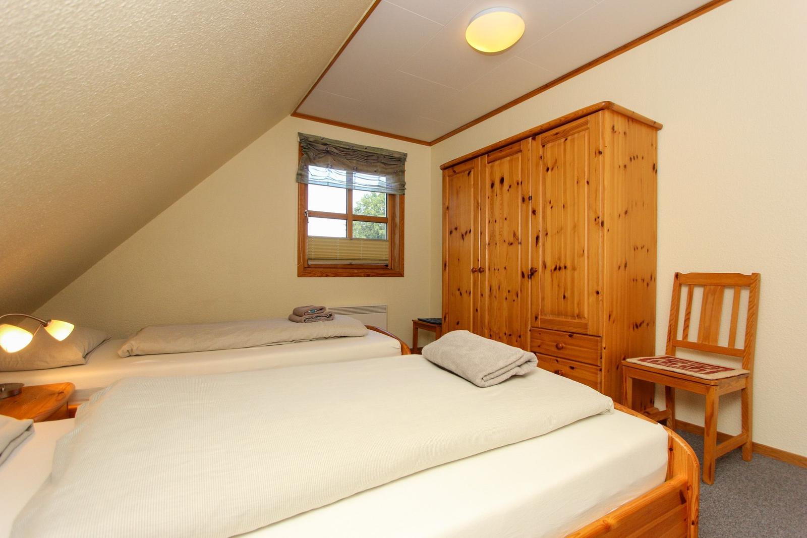 Zwei Betten, Holzschrank und Stuhl im Dachzimmer mit Fenster und Deckenleuchte.