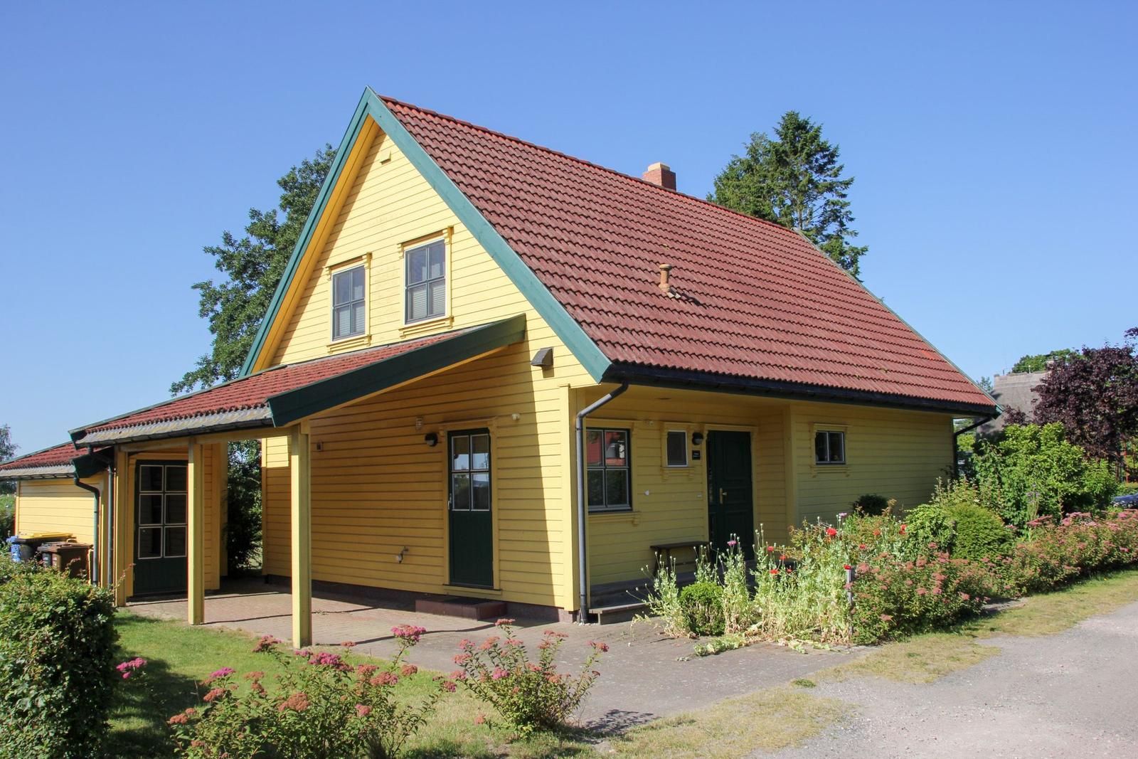 Gelbes Haus mit rotem Dach und grünen Fensterrahmen, umgeben von Grün und Blumen.