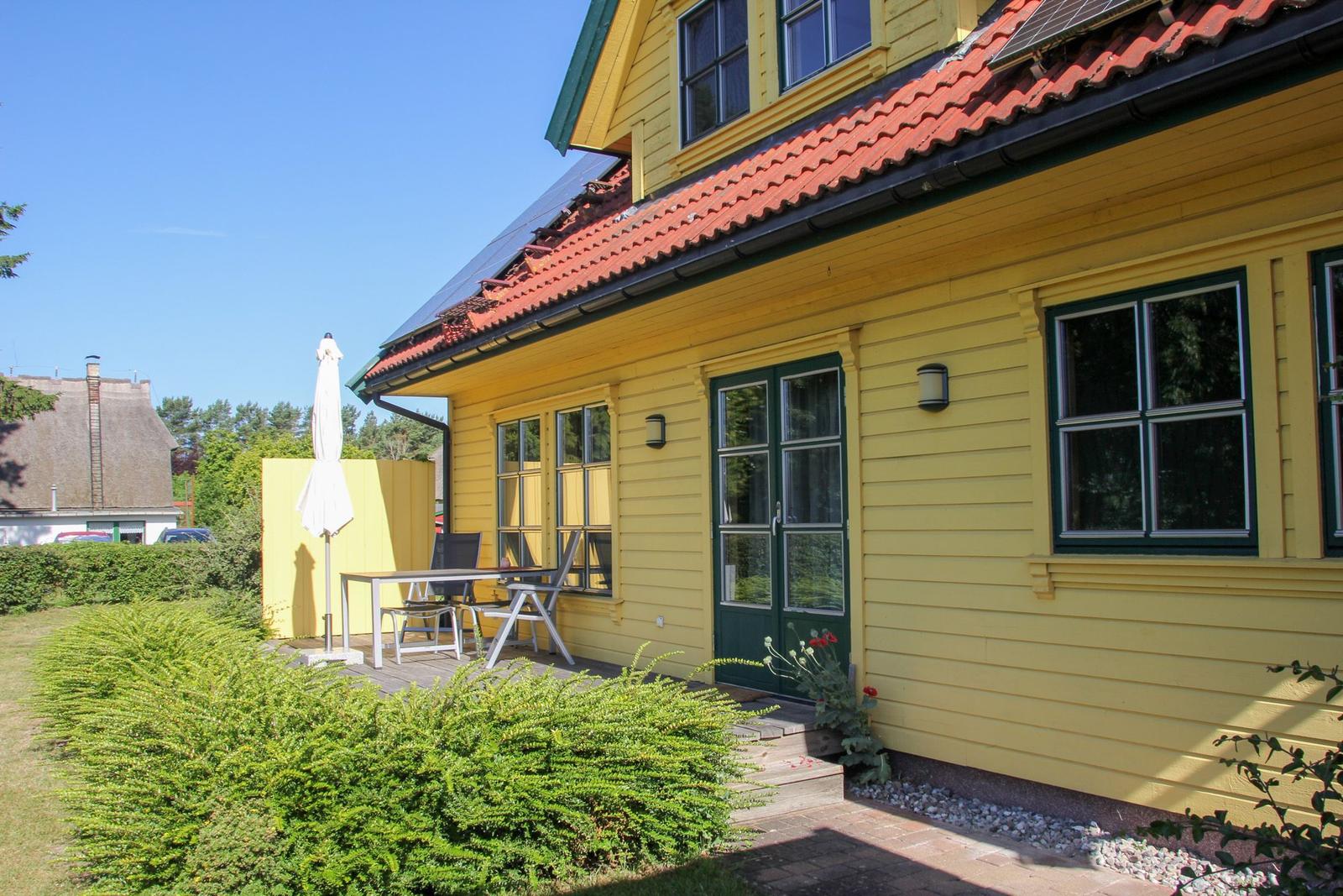 Gelbes Haus mit Terrasse, Tisch, Stühlen und Sonnenschirm. Roter Ziegeldach und grüne Fensterläden.
