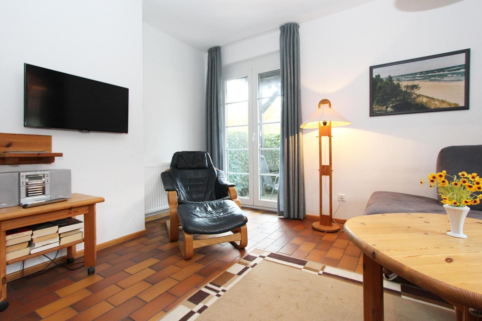 Wohnzimmer mit Fernseher, Sessel, Tisch und Fenster. Holzboden und Wandbild.