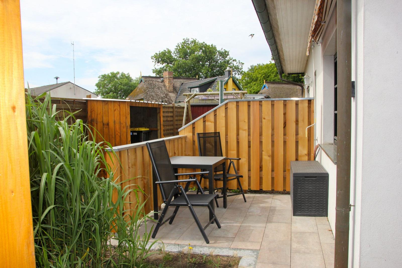 Terrasse mit Tisch und zwei Stühlen, umgeben von Holzgeländer und Grünflächen.