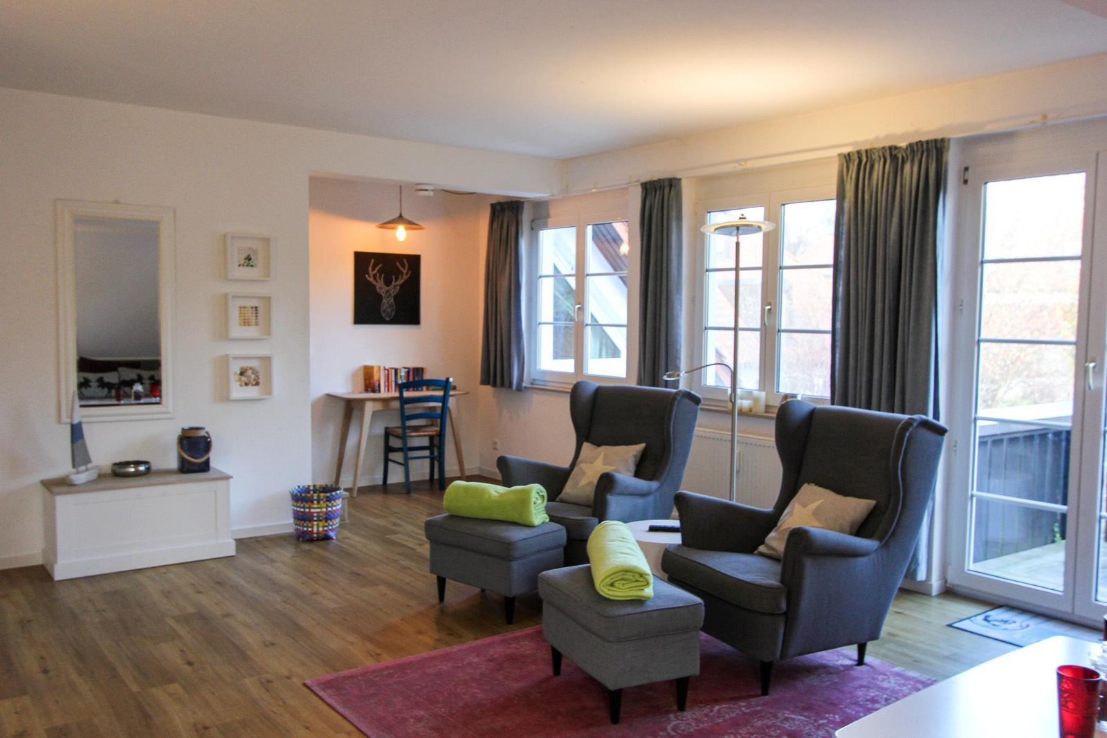 Wohnzimmer mit zwei Sesseln, Fußmatten und Fenstern. Hintergrund zeigt Arbeitsplatz.