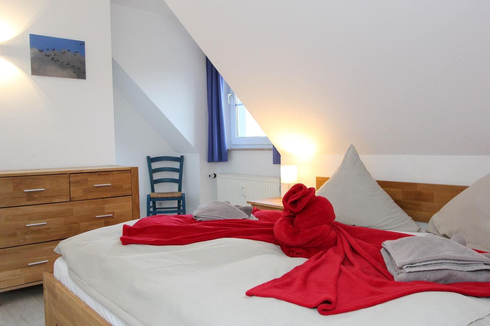 Schlafzimmer mit Dachschräge, Bett mit roter Decke, Holzkommode und blauer Stuhlecke.