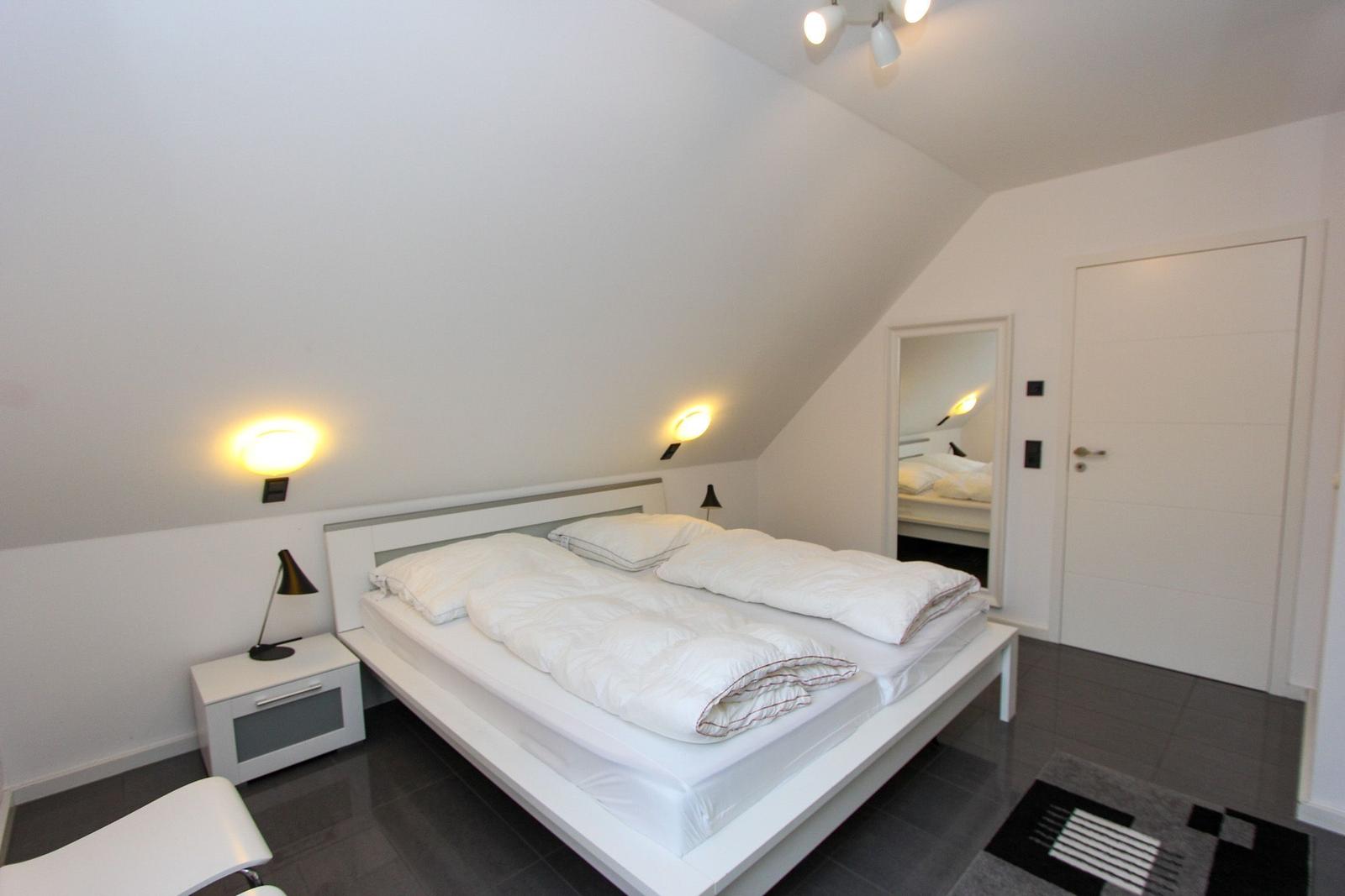 Schlafzimmer mit Doppelbett, Nachttisch und Spiegel. Dachneigung und dunkler Boden.