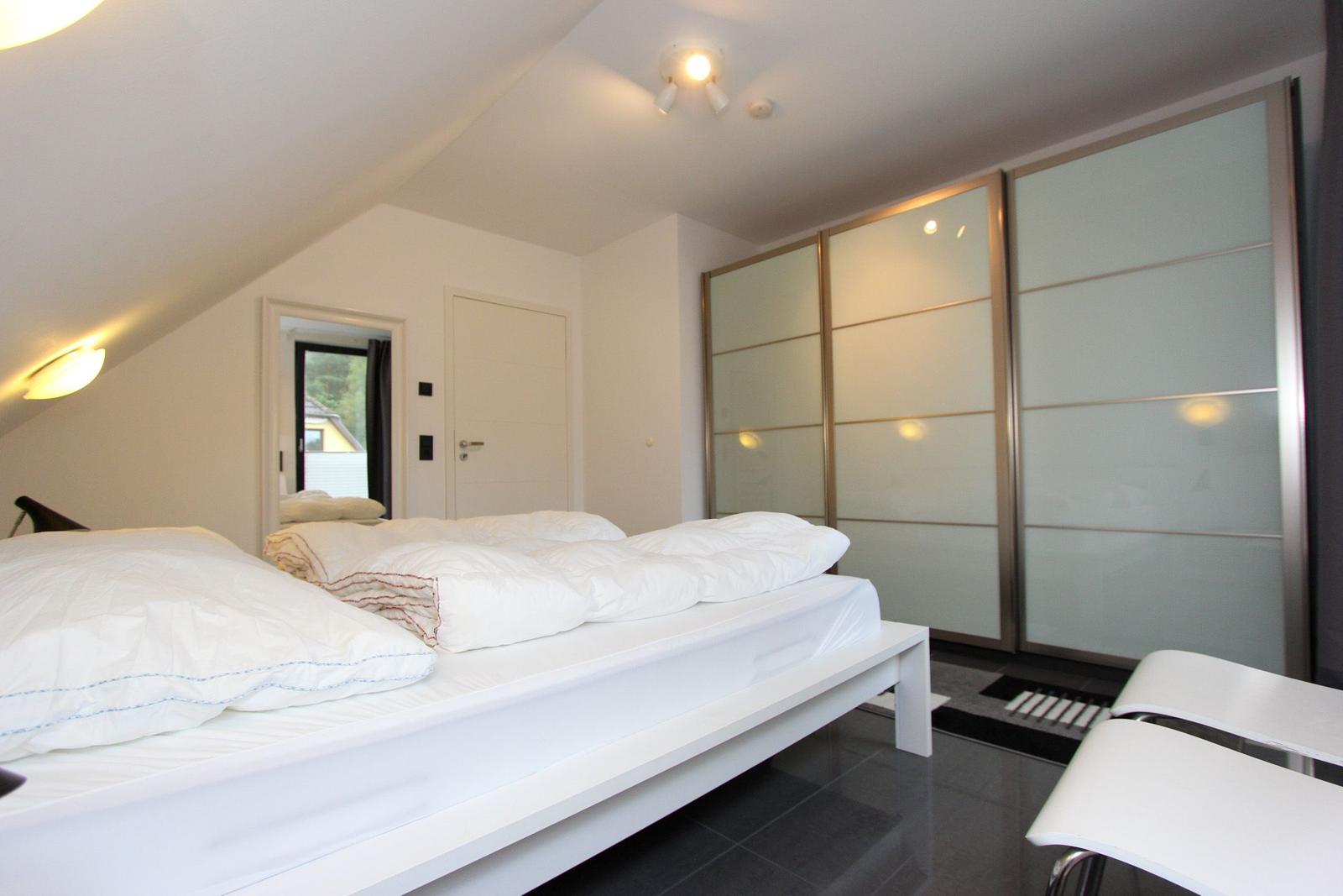 Schlafzimmer mit weißem Bett, Glaswand-Schrank und Dachschrägen.