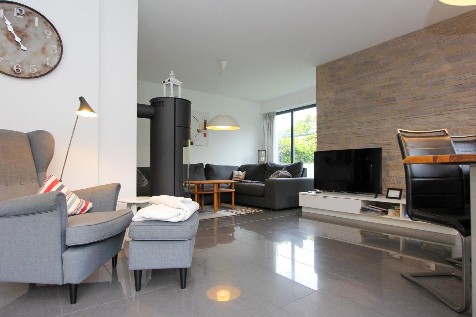 Wohnzimmer mit Sofa, Fernseher und Holzwand. Links Sessel und Stehlampe. Boden glänzend poliert.