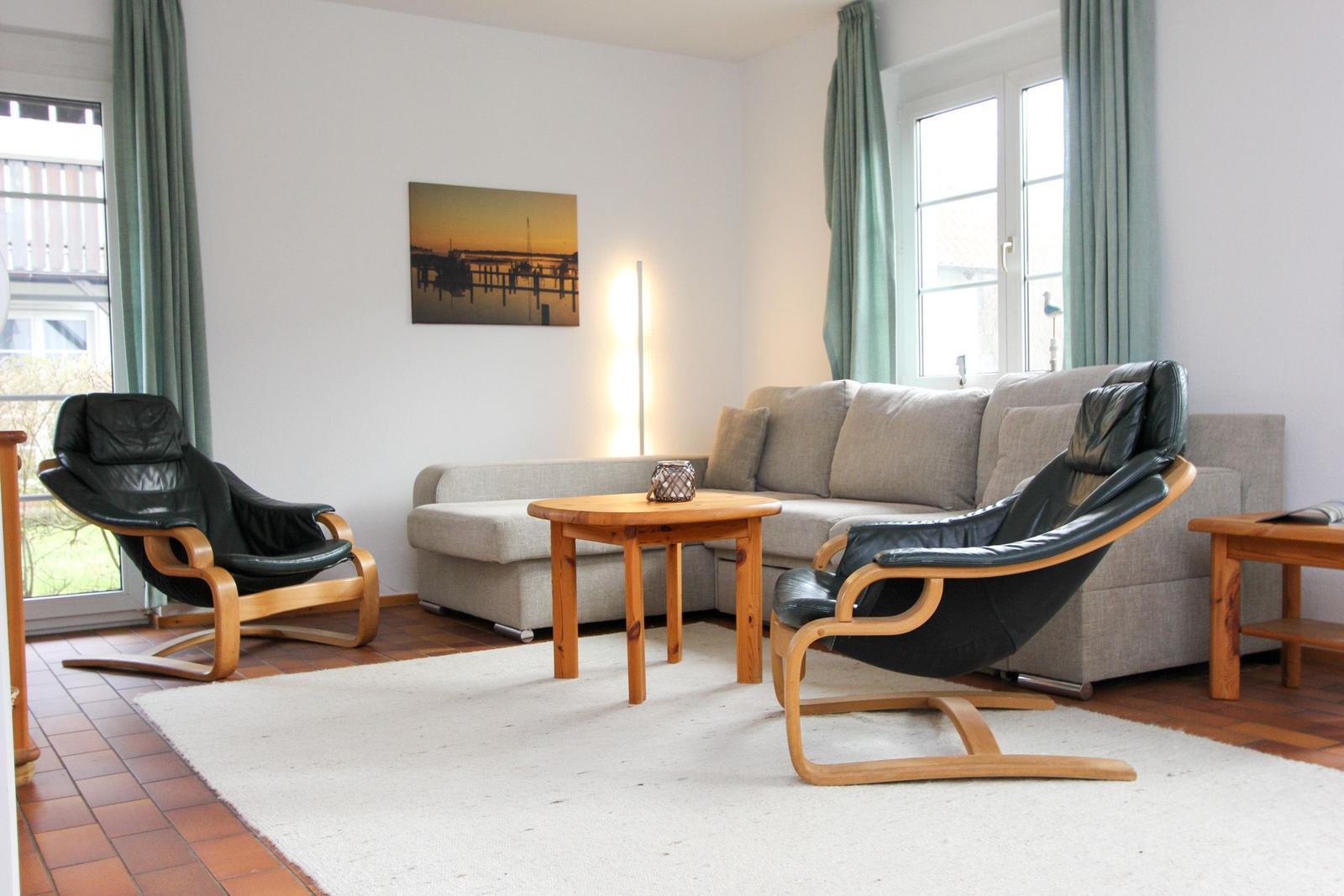 Wohnzimmer mit Sofa, zwei schwarzen Ledersessel und Holztisch. Fenster mit grünen Vorhängen.