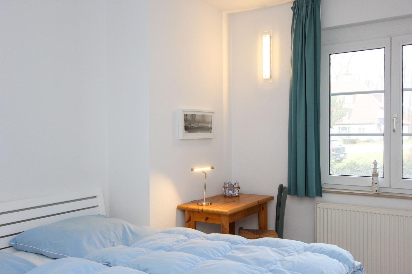 Schlafzimmer mit Bett, Tisch, Stuhl und Fenster. An der Wand hängt ein Bild und eine Lampe.