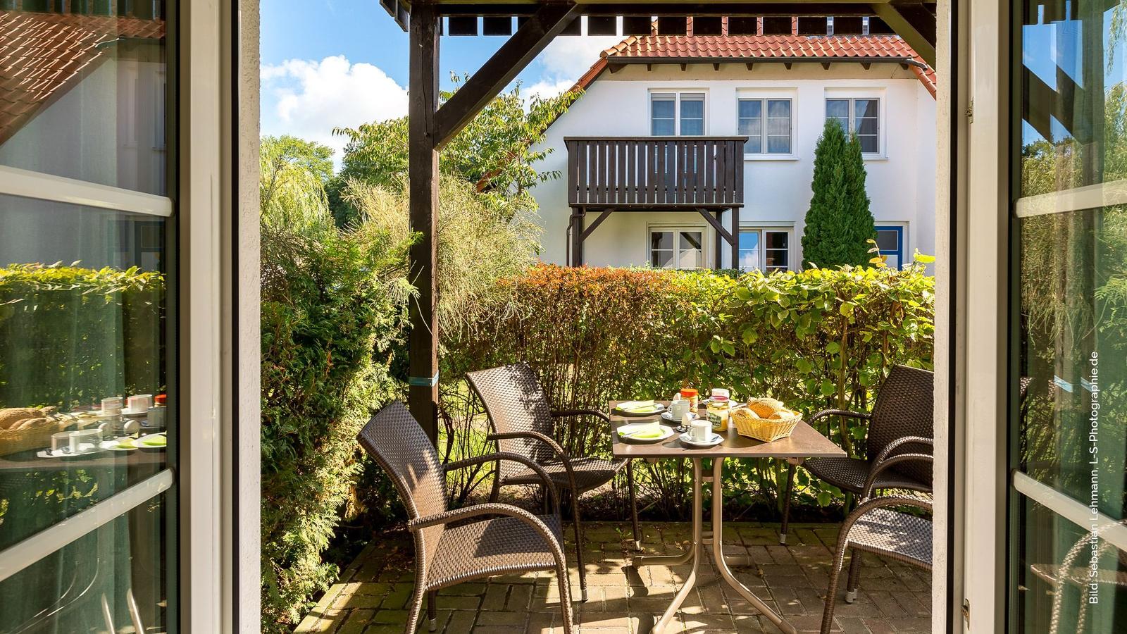 Terrasse mit Tisch und Stühlen, umgeben von grünen Hecken und einem weißen Haus im Hintergrund.
