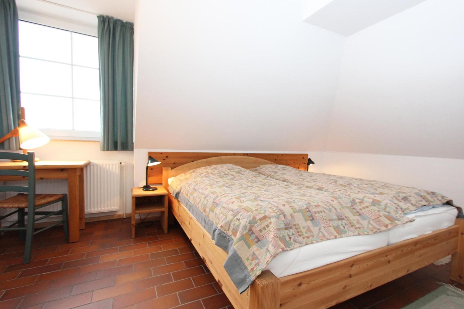Schlafzimmer mit Holzbett, Schreibtisch und Fenster. Boden aus Terrakottafliesen.