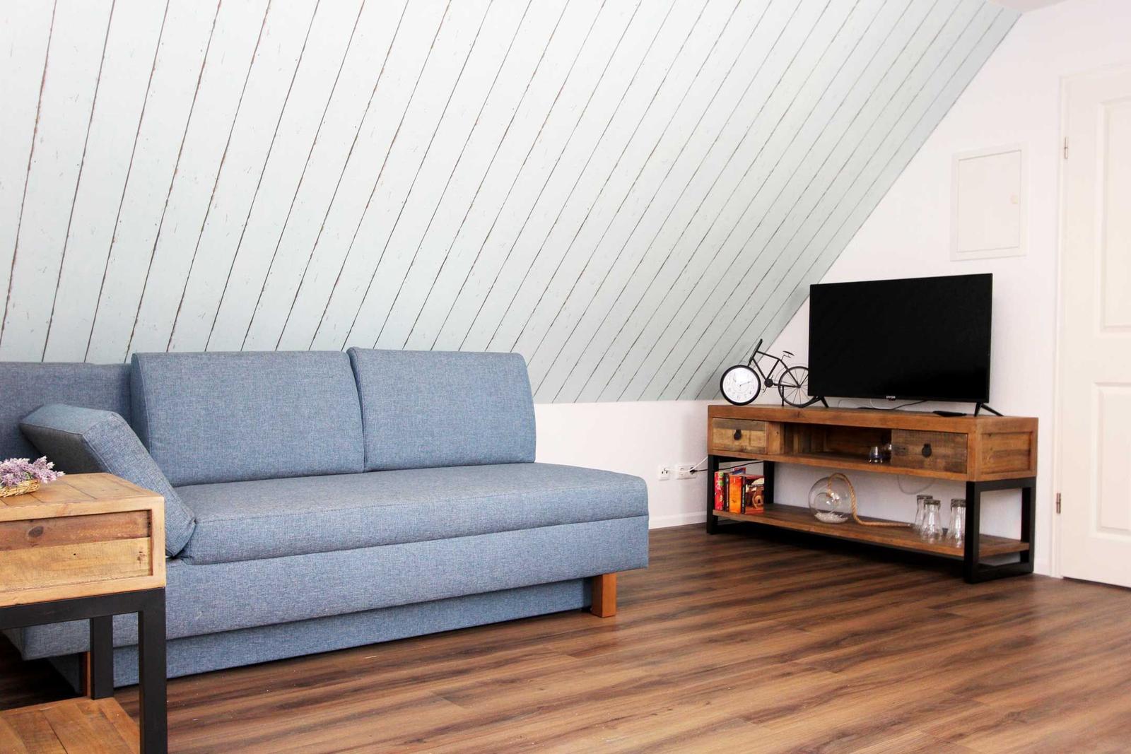 Ein Wohnzimmer mit blauem Sofa, Holzfußboden und schräger Holzwand. Fernseher auf Holzregal.