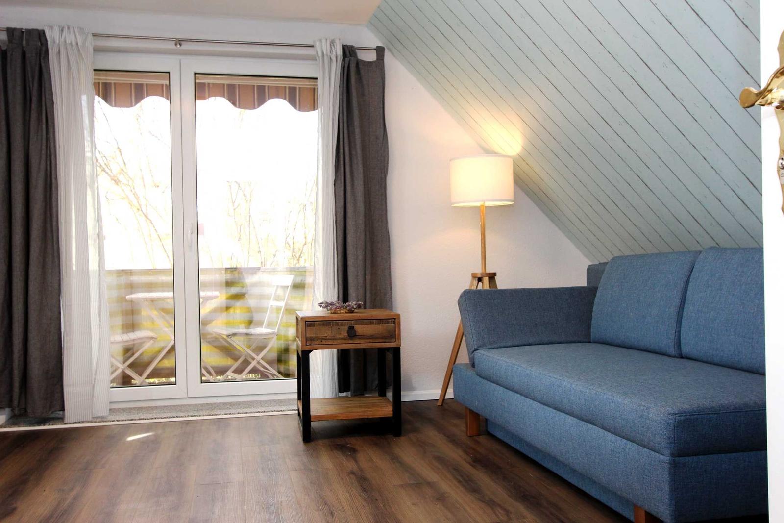 Ein Wohnzimmer mit blauem Sofa, Holzboden und Dachschrägen. Vor dem Fenster befindet sich eine Terrasse.