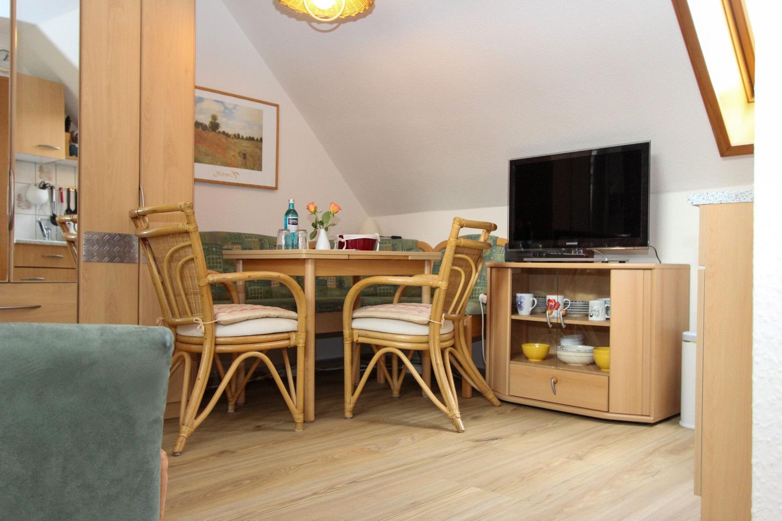 Esszimmer mit Holztisch, Stühlen und Fernseher. Wandbild und Küchenbereich sichtbar.
