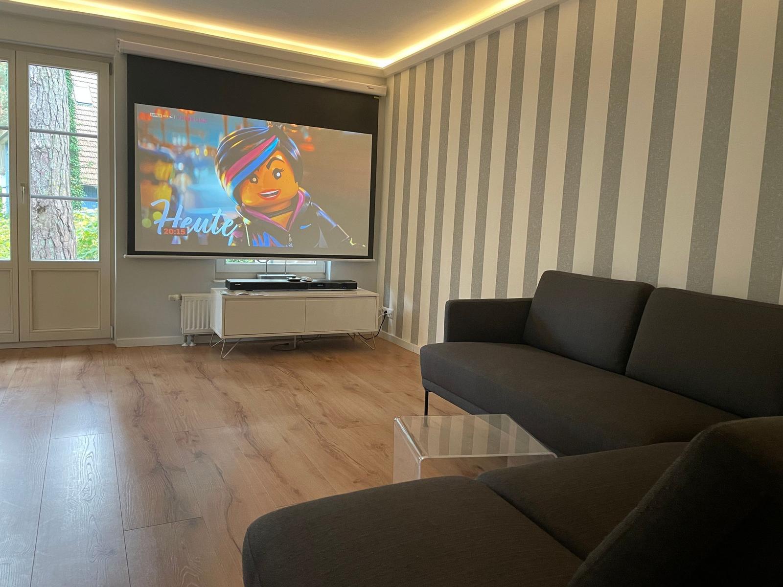 Ein Wohnzimmer mit einem Sofa, einem Glas-Tisch und einer Projektionswand.