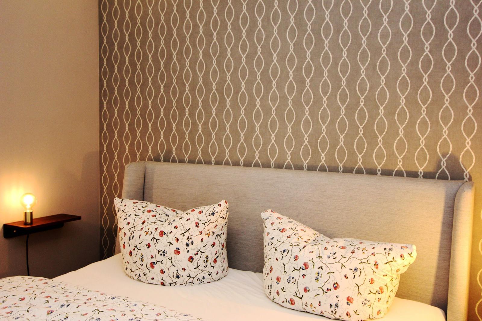 Doppelbett mit floralen Kissen und Tapete mit Muster.