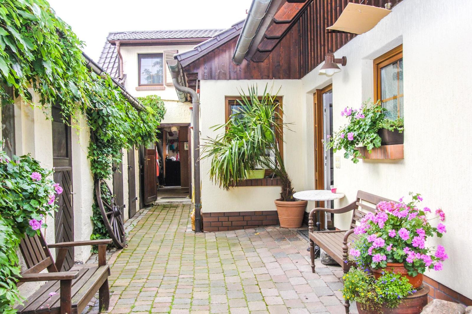 Hinterhof mit Steinplatten, Pflanzen und Holzbank. Eingangstür und Fenster sichtbar.