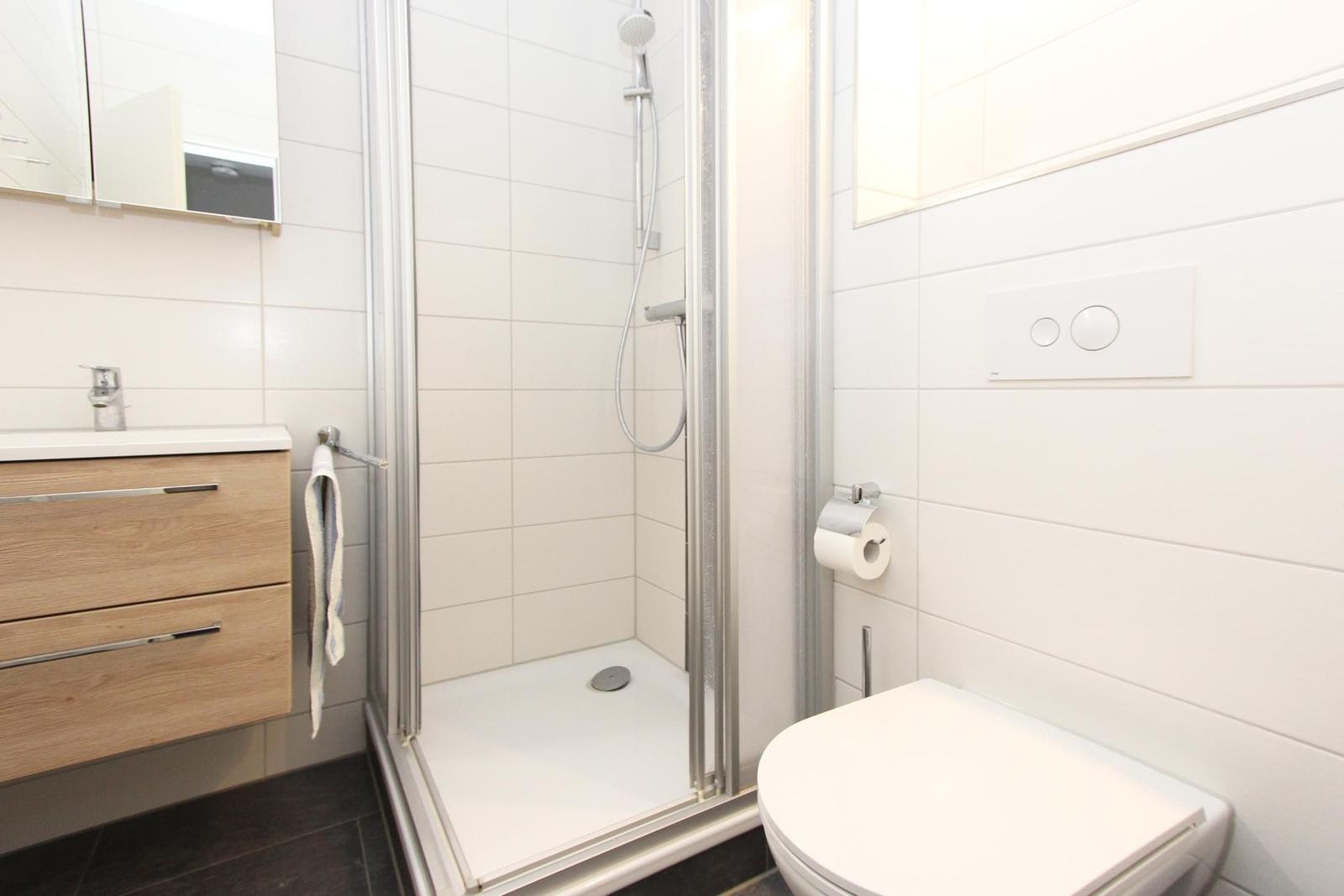 Badezimmer mit Dusche, Waschbecken und Toilette. Weiße Fliesen und Holzwaschtisch.
