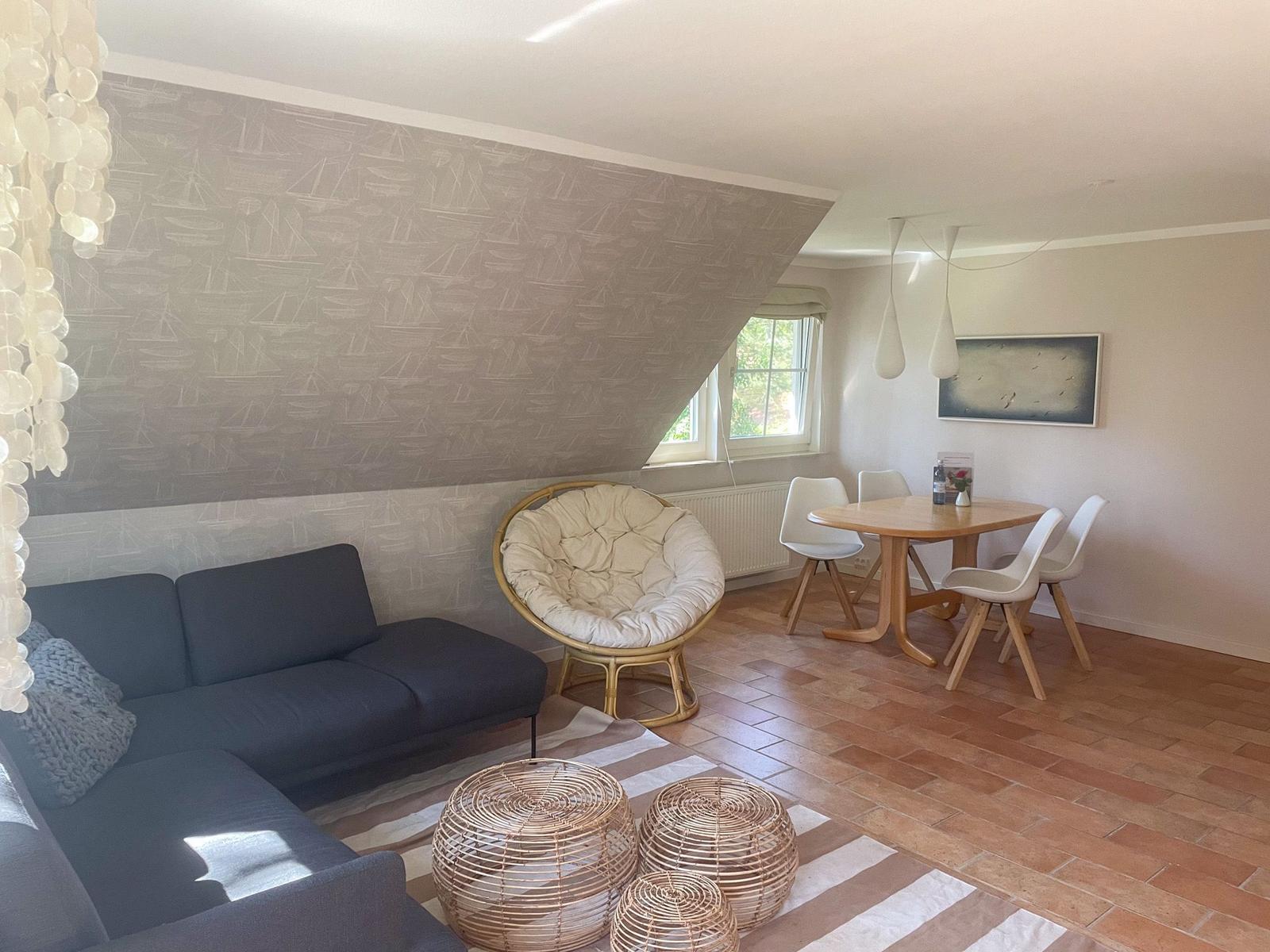 Wohn- und Essbereich mit Sofa, Rattanmöbeln und Holztisch. Wand mit Segelyacht-Motiv.