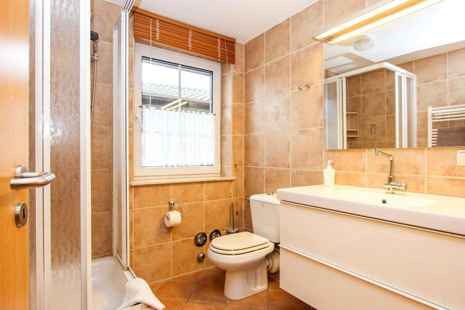 Badezimmer mit Dusche, Toilette und Waschbecken. Fenster mit Jalousie und Spiegel.