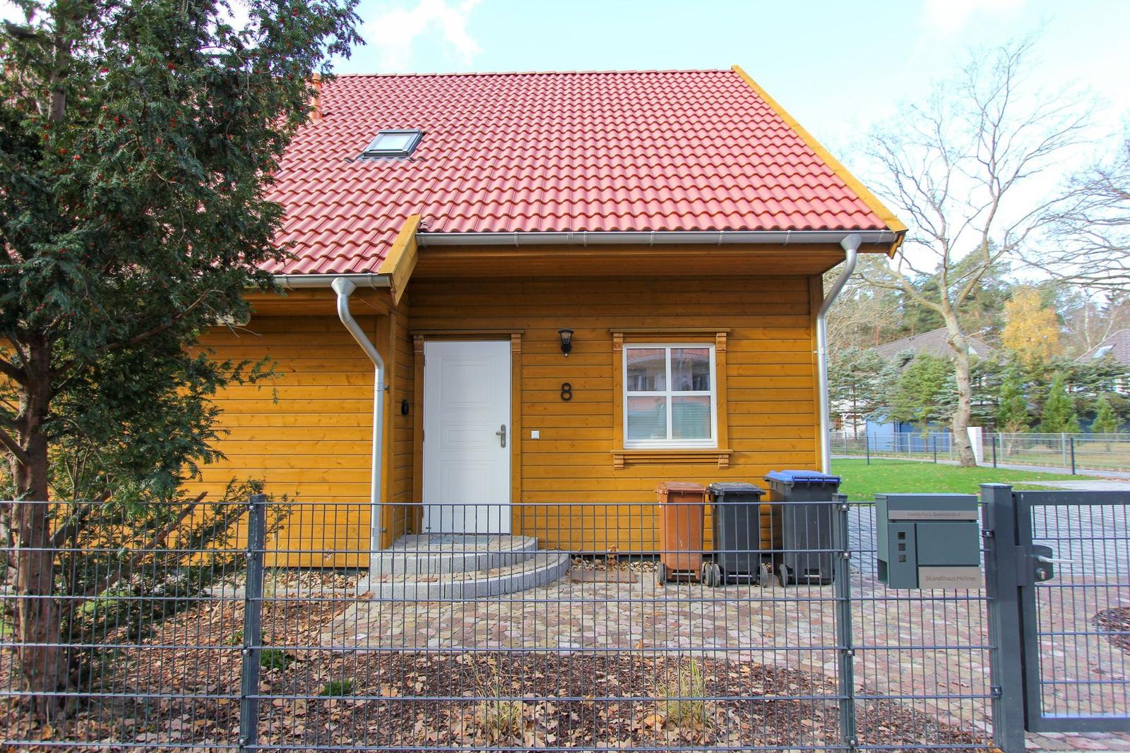 Holzhaus mit rotem Dach, weiße Tür und Fenster, Hof mit Zaun und Mülltonnen.