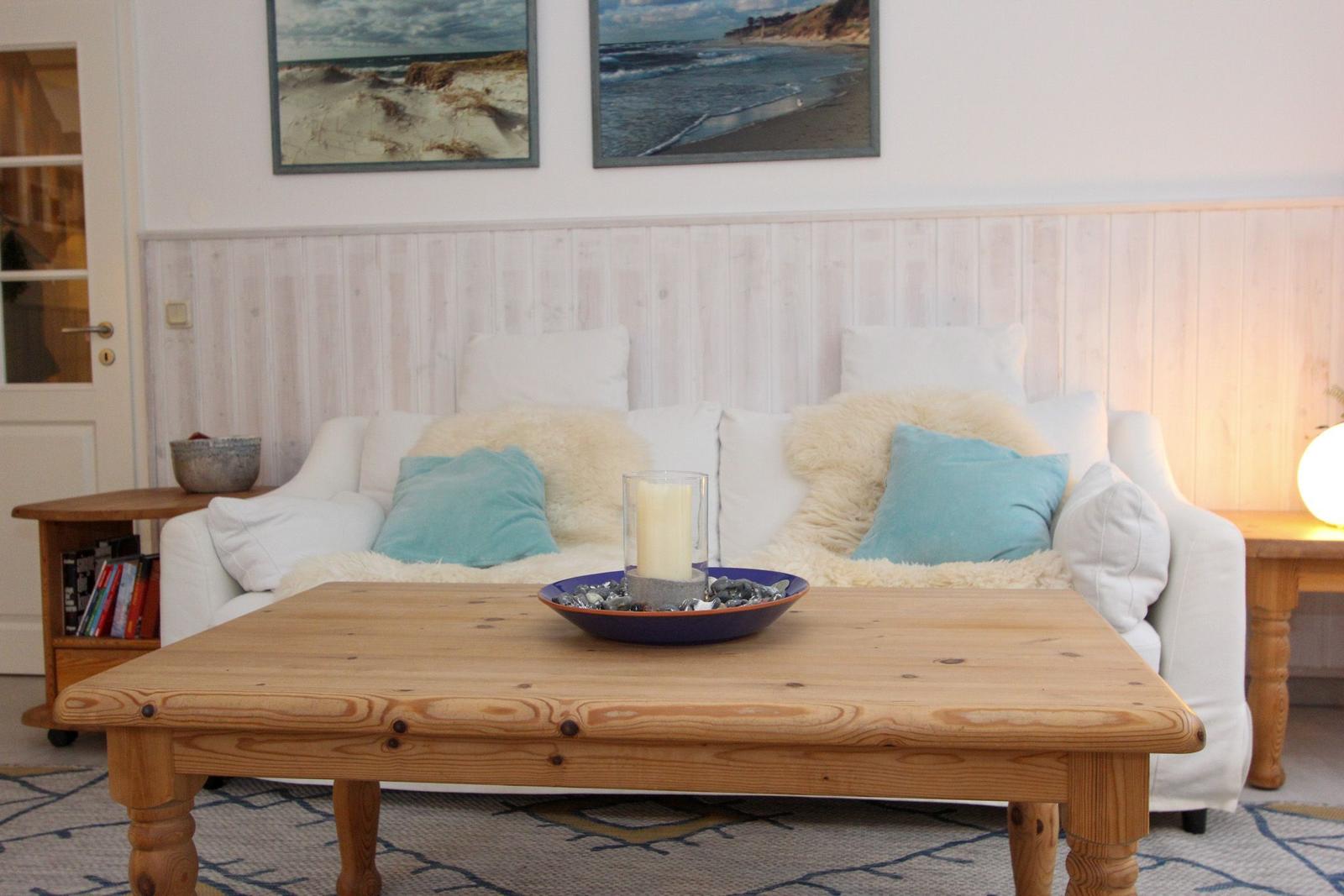 Ein Wohnzimmer mit weißem Sofa, Holztisch und Strandmotiven an der Wand.