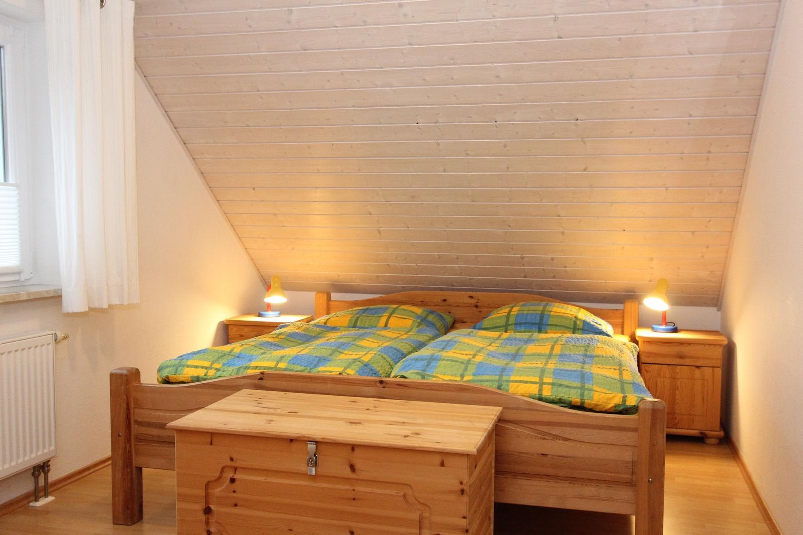 Doppelbett mit grün-gelbem Bettzeug, zwei Nachttische mit Lampen, Holzdecke und Fenster mit Vorhang.