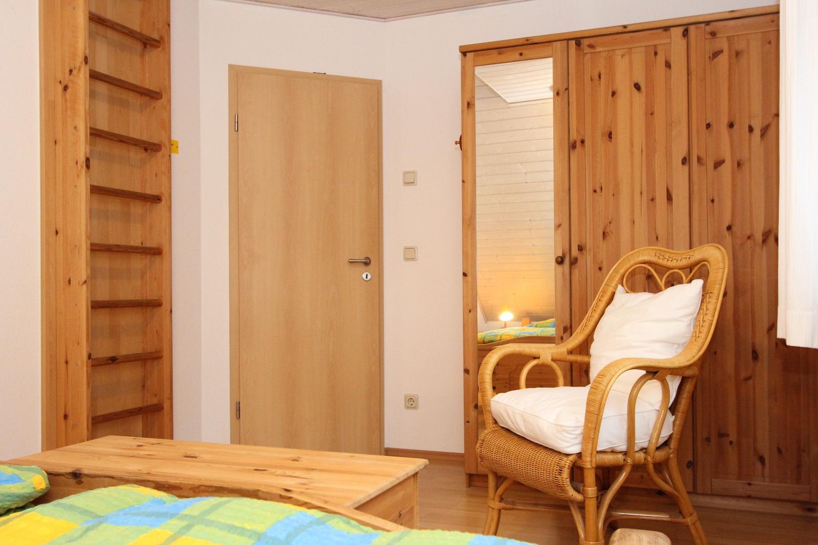 Schlafzimmer mit Holzbett, Stuhl und Kleiderschrank. Tür und Wand sind sichtbar.
