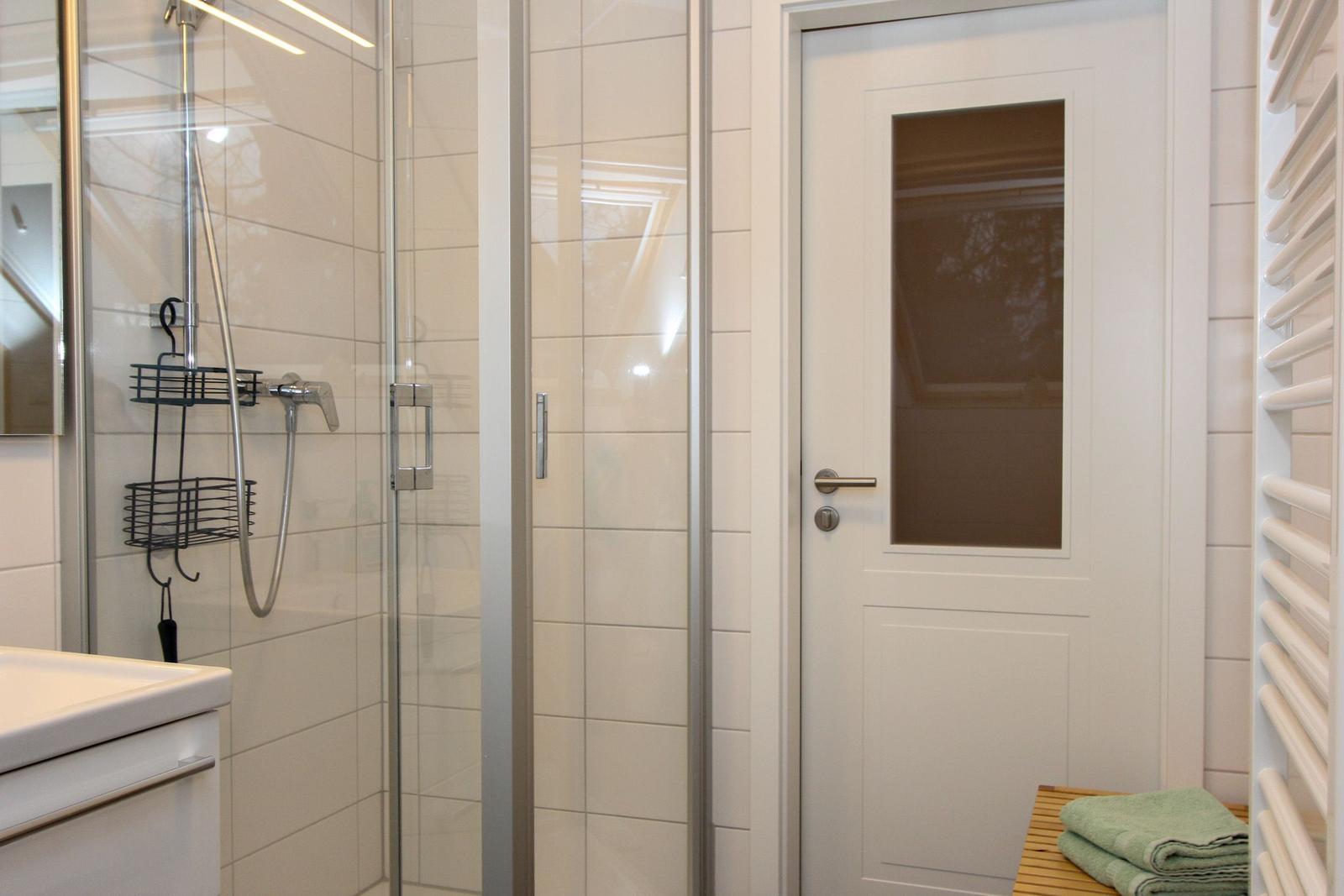 Dusche mit Glaswand, weiße Fliesen und Handbrause. Tür mit Glasfenster und Handtuch auf Badebank.