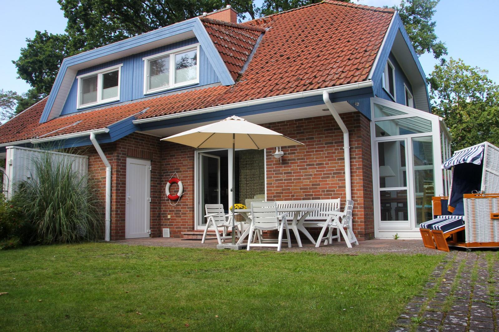 Haus mit Terrasse, Tisch, Stühlen und Sonnenschirm. Garten und Strandkorb sind vorhanden.