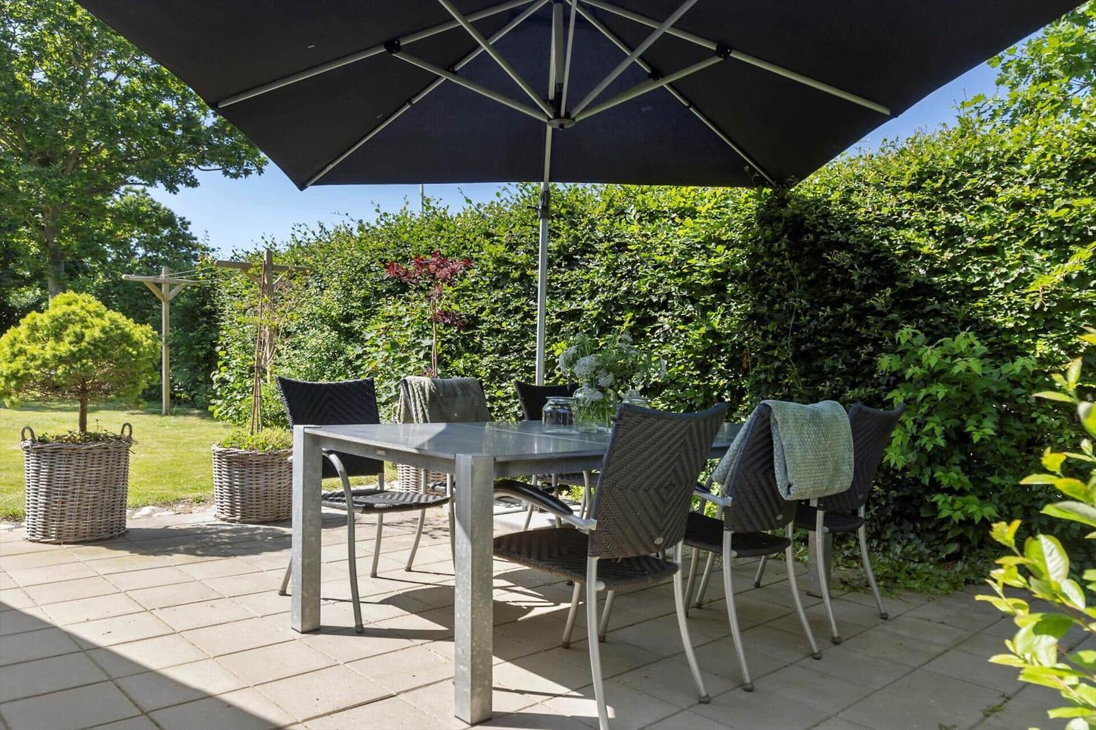Terrasse mit Tisch, Stühlen und Sonnenschutz. Hintergrund: grüne Hecke und Garten.