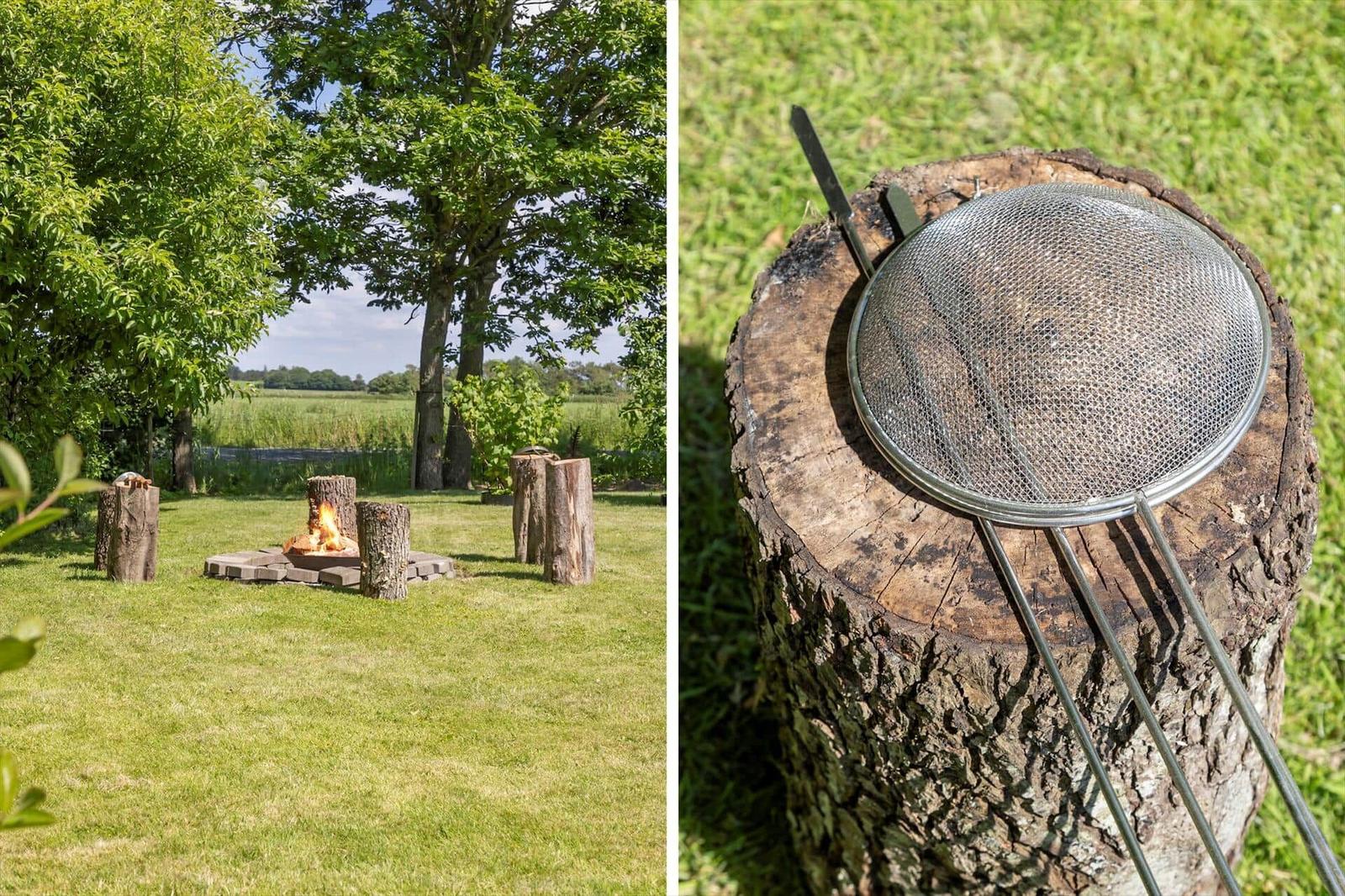 Außenbereich mit Grillplatz und Holzstumpf als Sitzgelegenheit. Feuer brennt im Grillbereich.